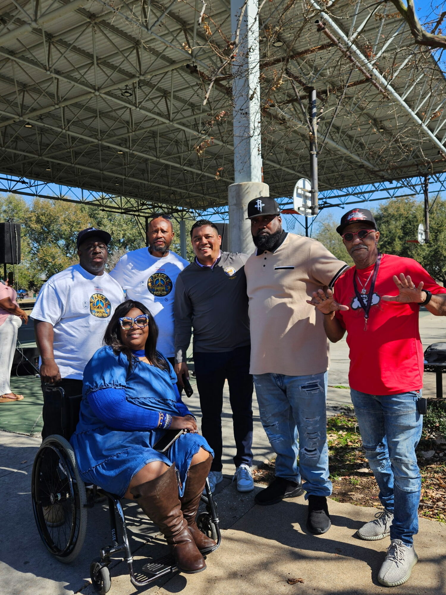 Group of six people outdoors under a large metal structure, some standing and one woman in a wheelchair, smiling.