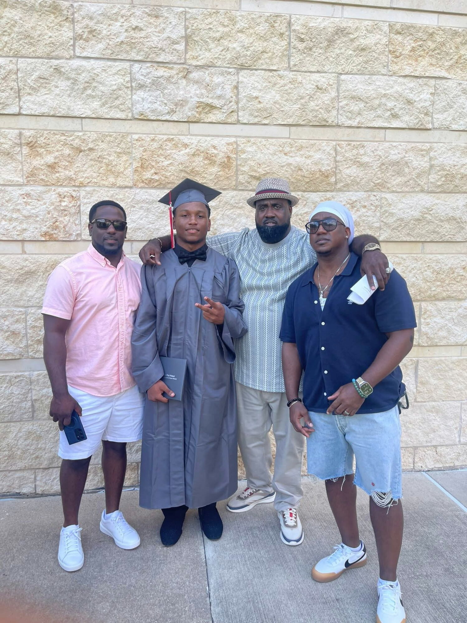 Four people standing together outdoors in front of a stone wall, one in graduation gown and cap, others casually dressed.