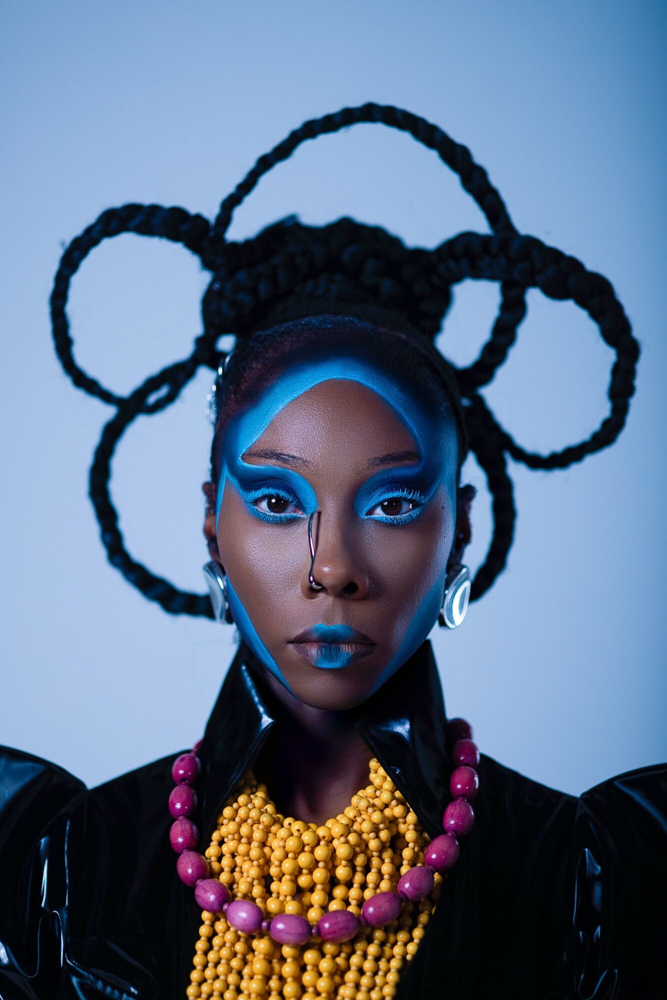 Portrait of a woman with elaborate braided hairstyle, vibrant blue makeup, and colorful jewelry against a blue background.
