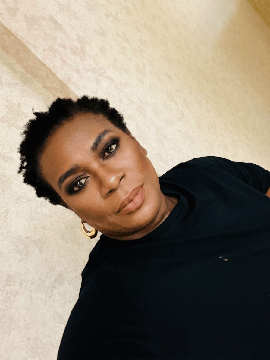 Woman with short curly hair, makeup, and gold hoop earrings, wearing a black top, looking at the camera.