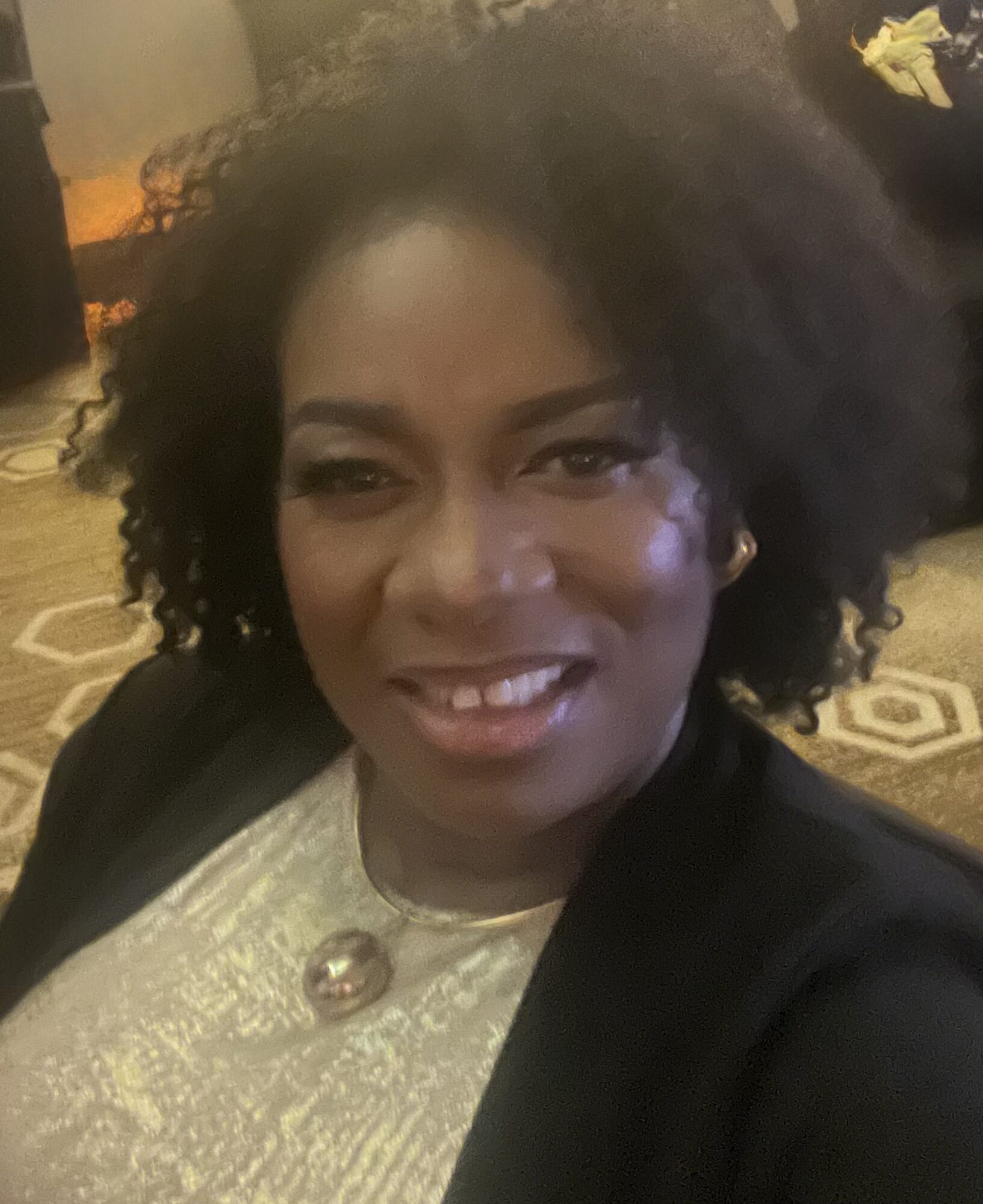 Woman with curly hair smiling, wearing a light-colored top and black jacket, indoors with patterned carpet.