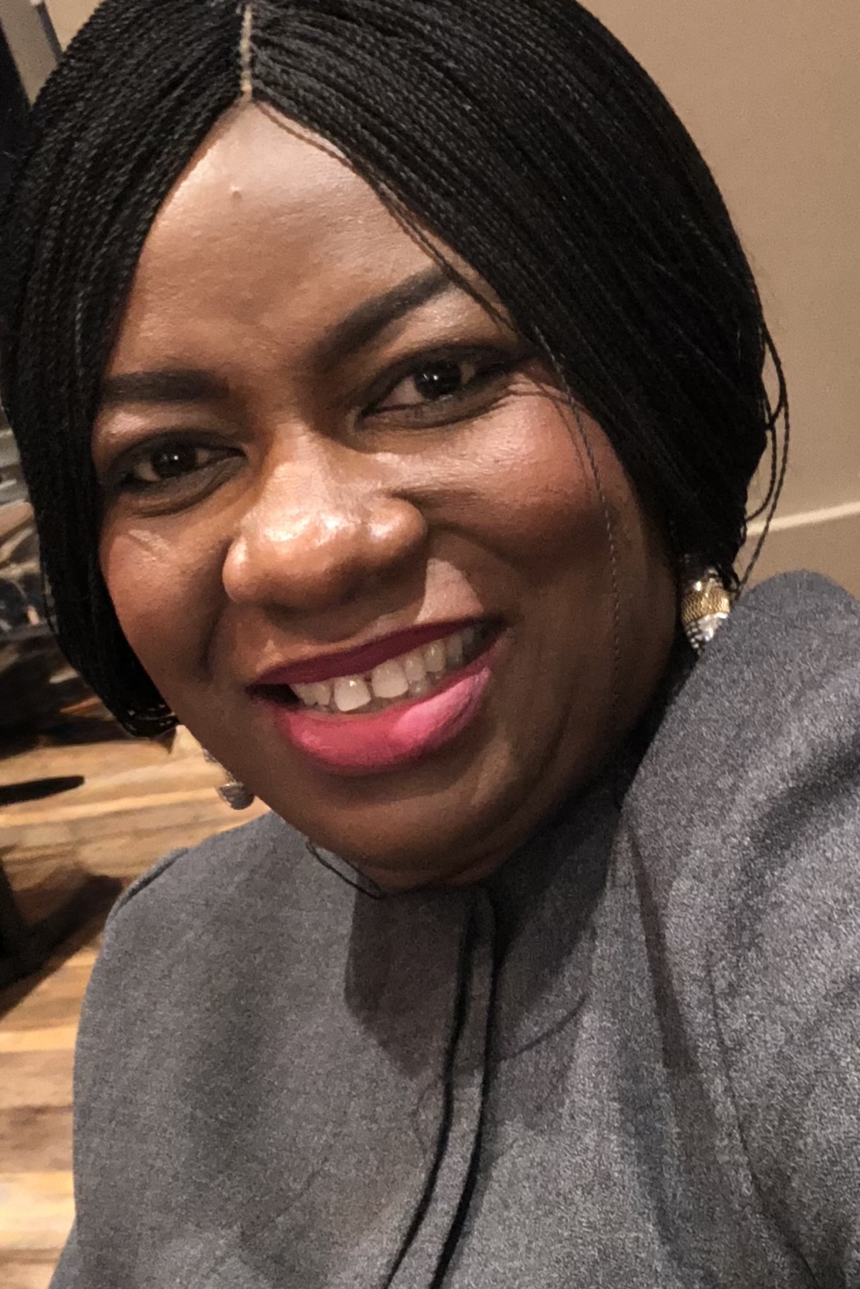 Close-up of a smiling woman with black braided hair, wearing a gray top and earrings.
