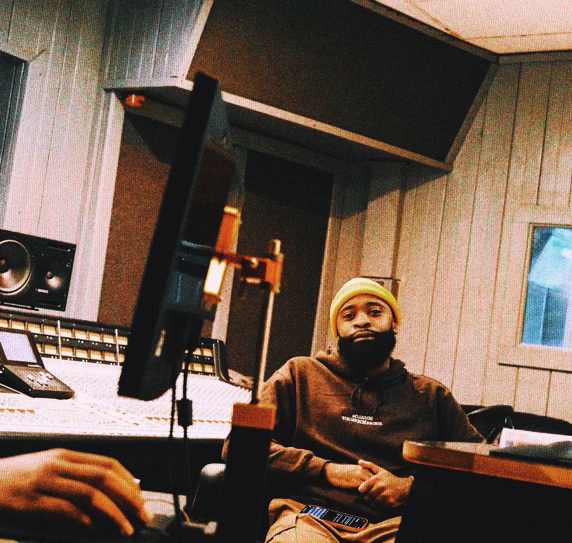 Person wearing a yellow beanie and brown jacket sitting in a recording studio with equipment around.