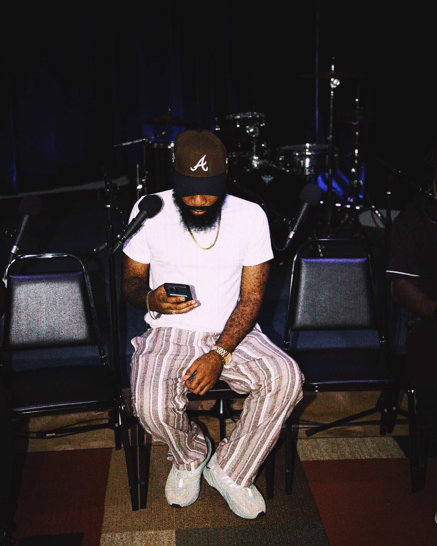 Person sitting on a chair, wearing a cap, white t-shirt, striped pants, and white shoes, looking at a phone.