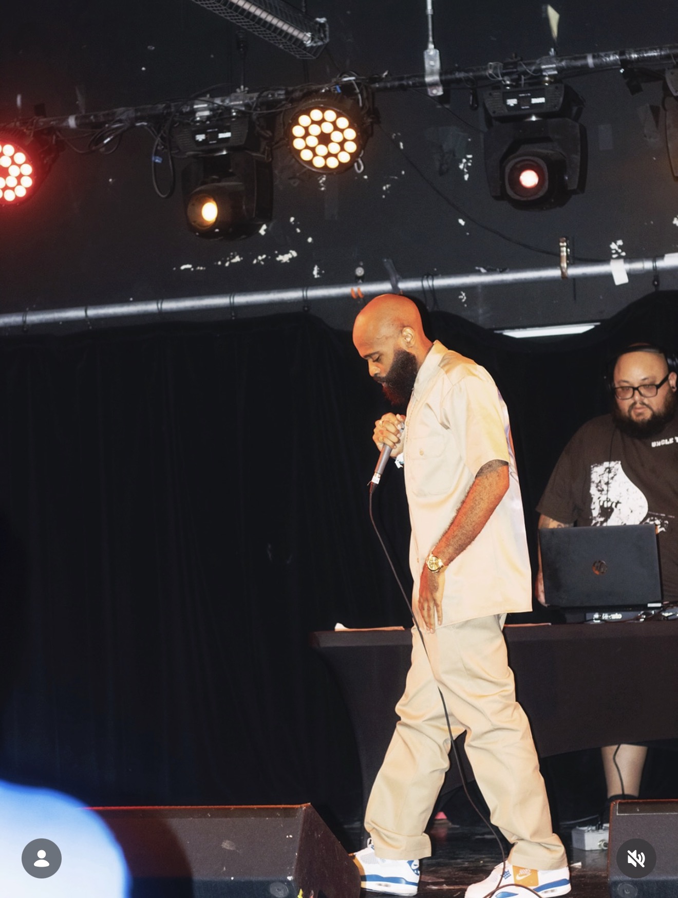 Performer with a microphone on stage, wearing light-colored clothing, with a DJ or music setup nearby, stage lights above.