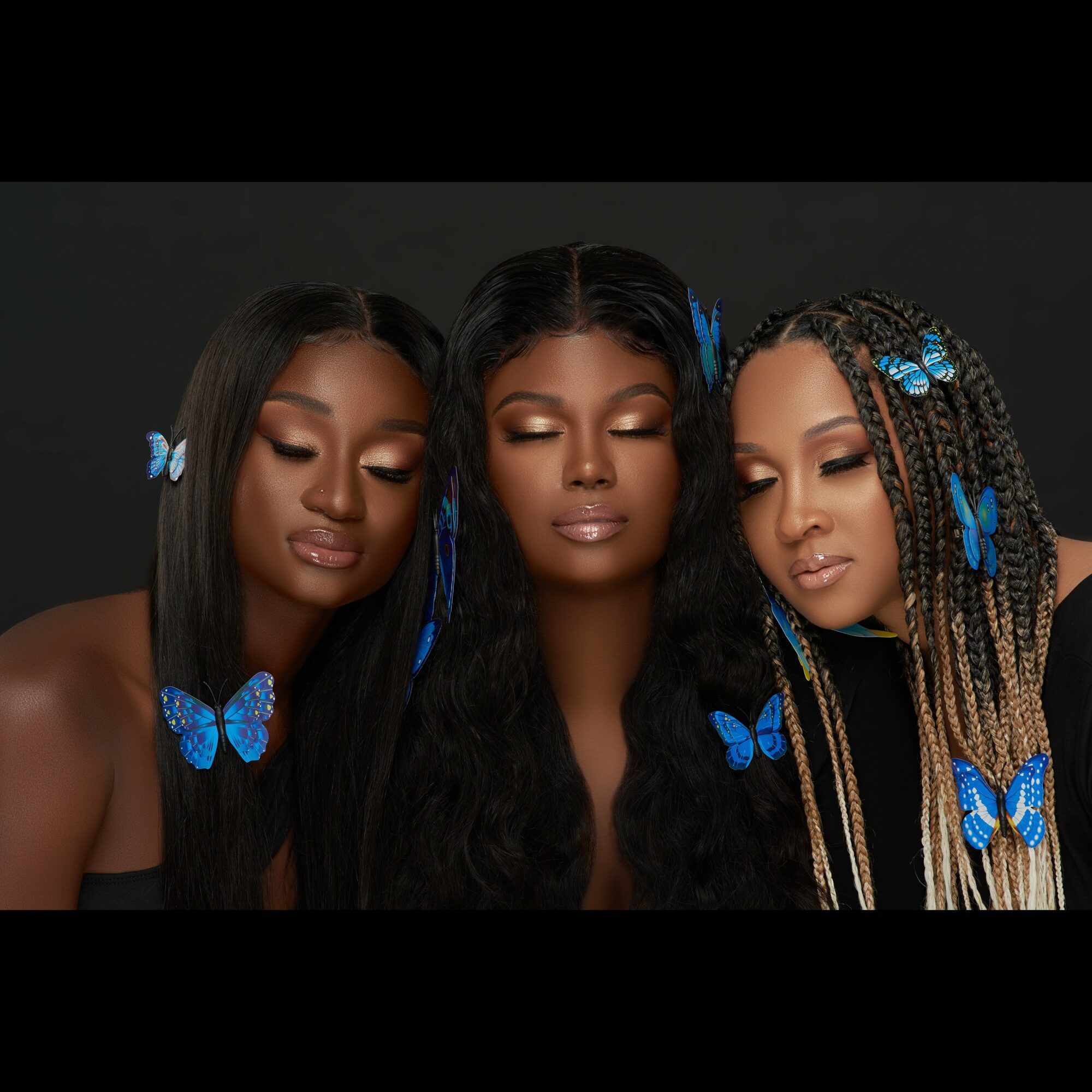 Three women with long hair and blue butterfly accessories pose against a black background.