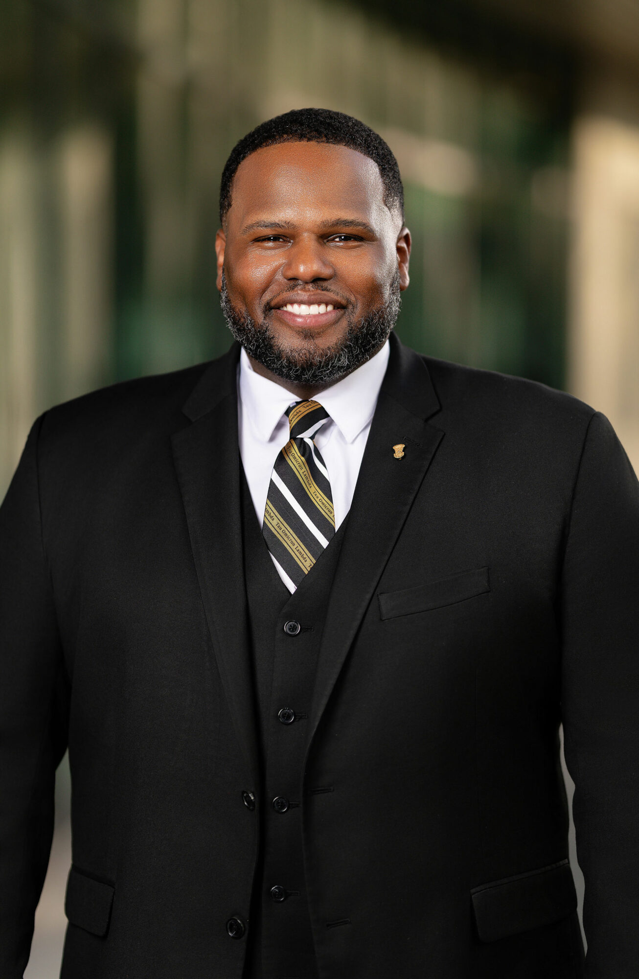 Man in a black suit and striped tie smiling outdoors with blurred background