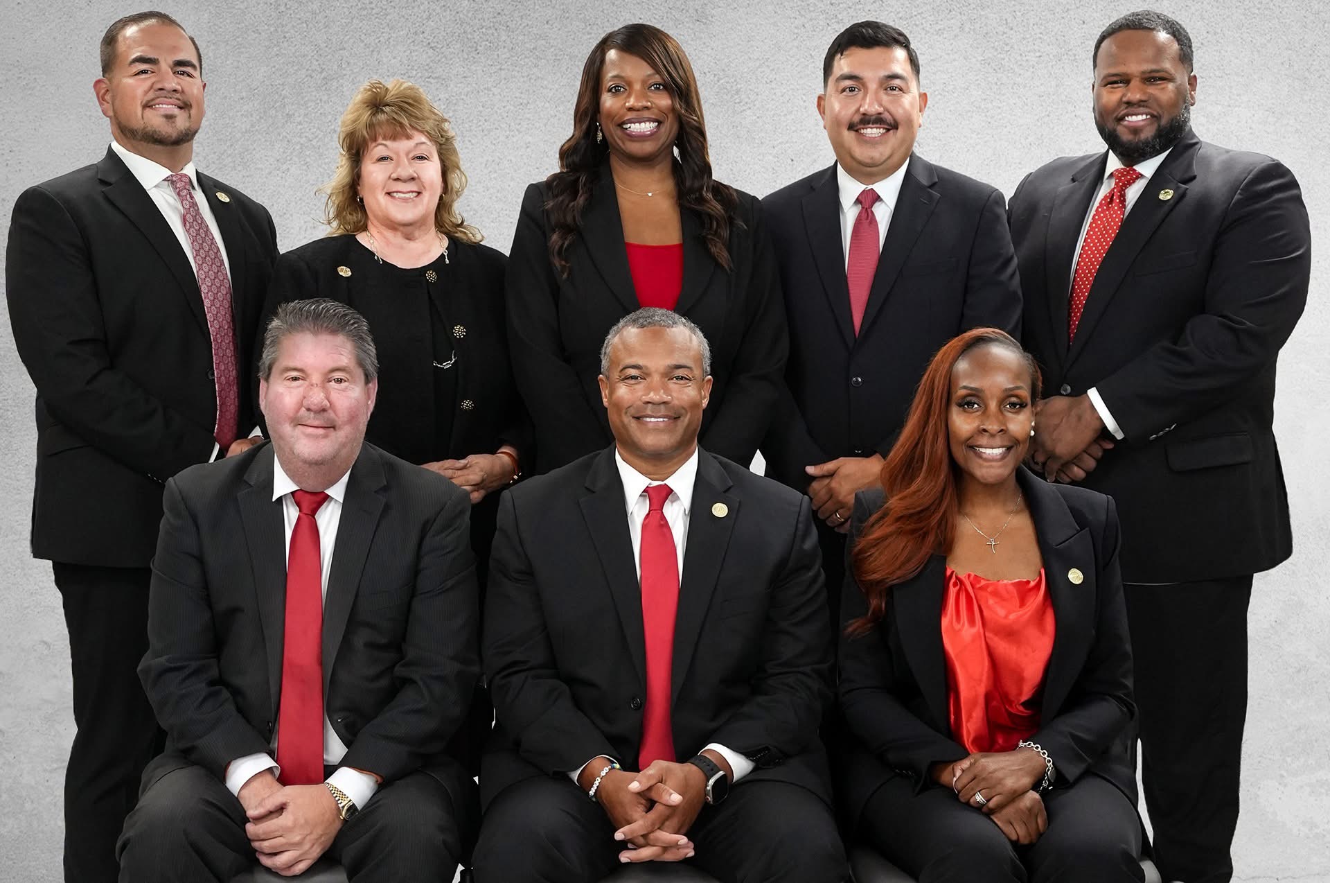 Group of eight diverse professionals in formal attire, smiling, standing and sitting against a gray background.