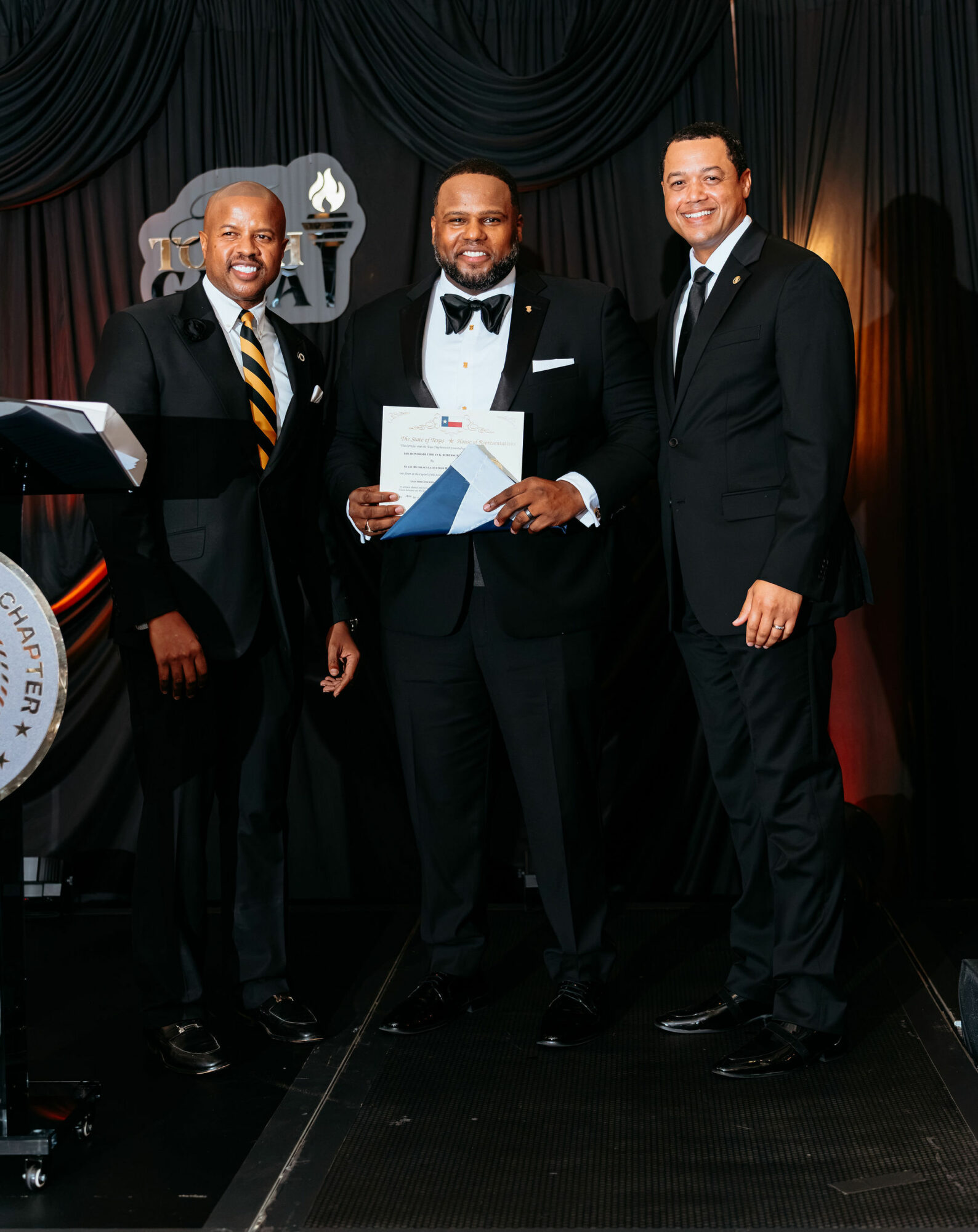 Three men in formal attire standing on stage, one holding a certificate, with a dark curtain background.