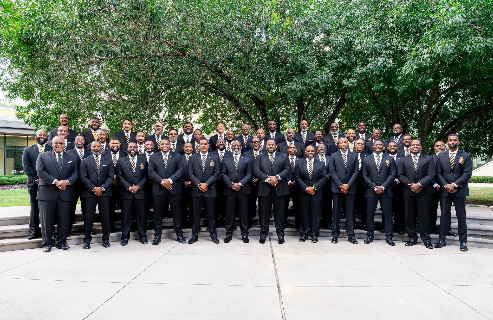 Group of men in suits standing outdoors on a sidewalk with trees in the background.