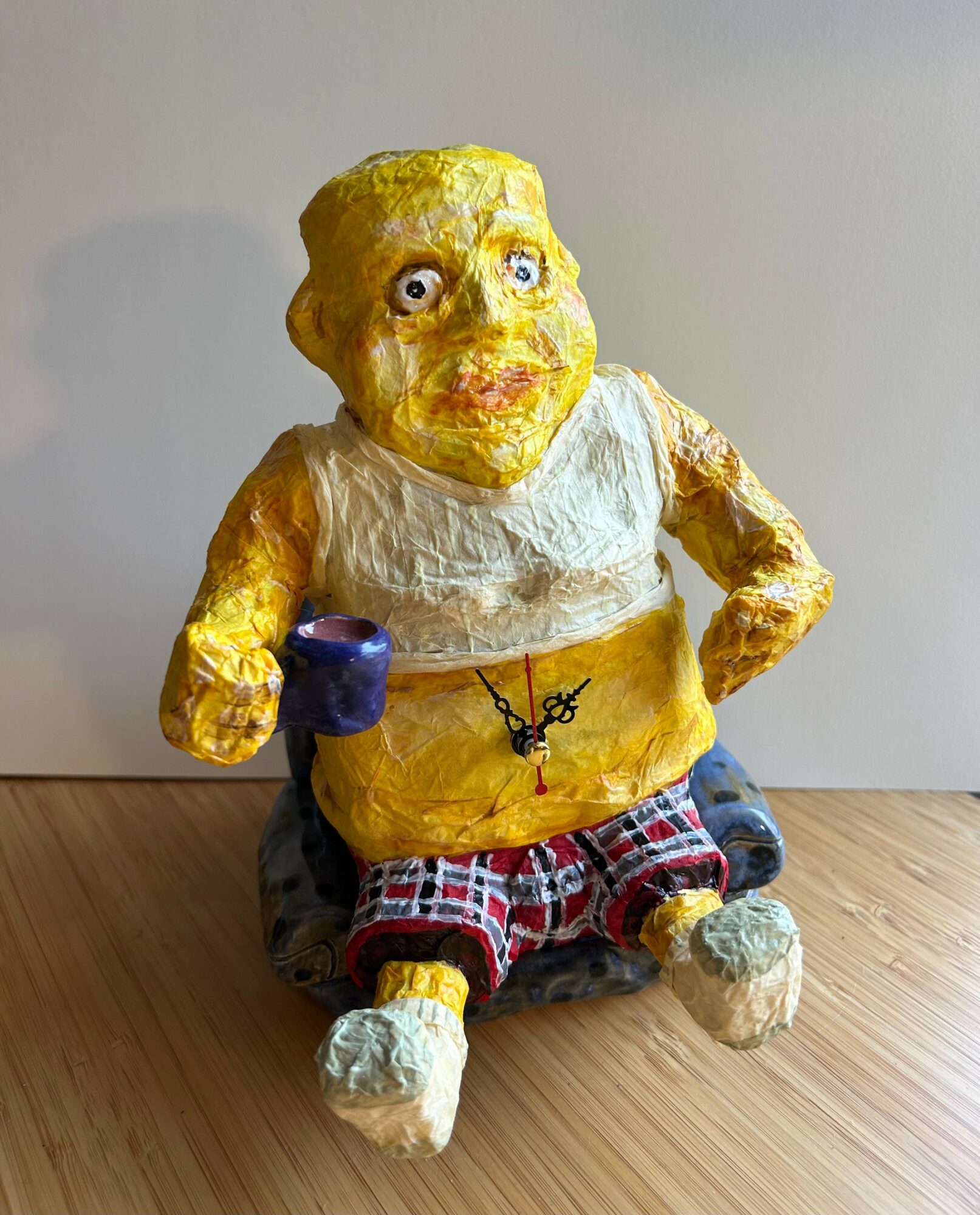Sculpture of a chubby person with yellow skin, wearing a white top and plaid shorts, sitting on a wooden surface.