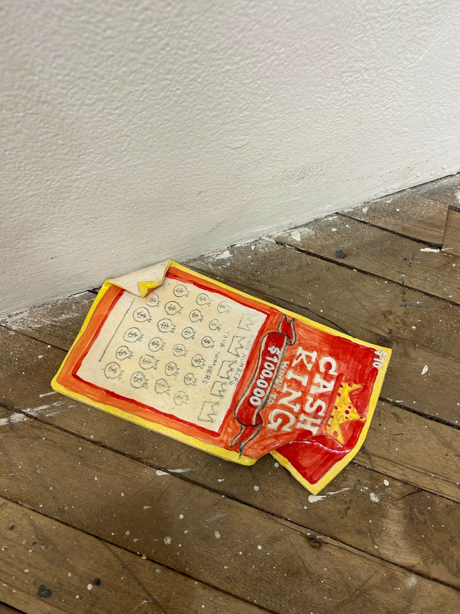 Crushed snack bag on wooden floor near wall, with visible text and symbols, some debris around it.