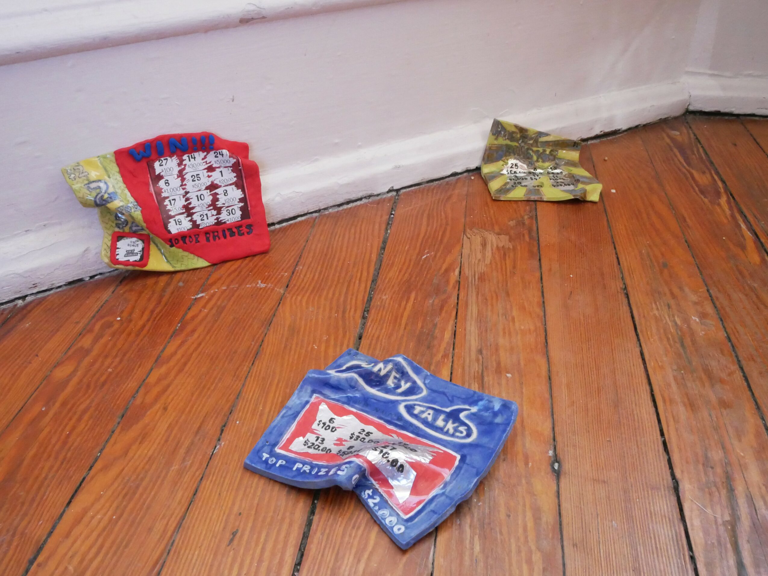Three crumpled snack bags on a wooden floor near a white wall, with one bag partially under the wall baseboard.