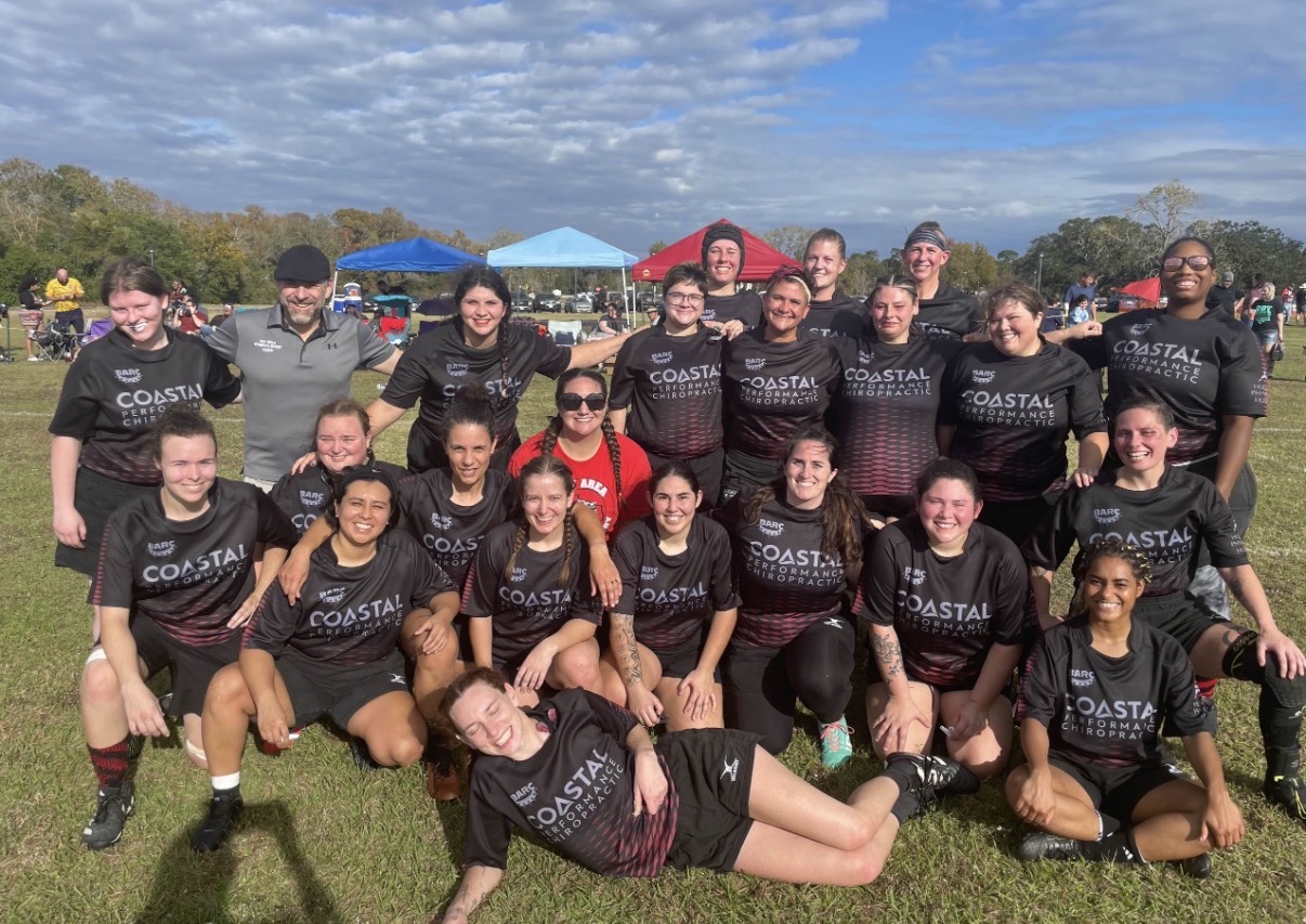 Group of women in black sports uniforms posing outdoors on grass, some standing and some kneeling, with tents and trees in background.