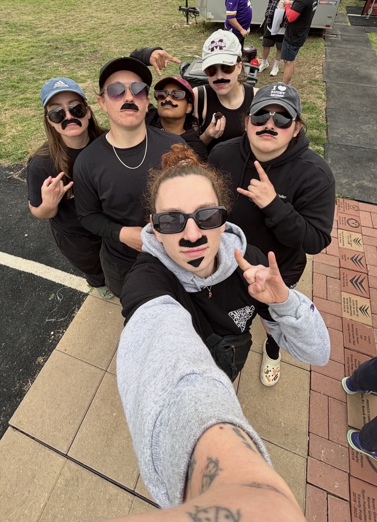 Group of six people outdoors, wearing sunglasses and fake mustaches, taking a selfie together on a sunny day.