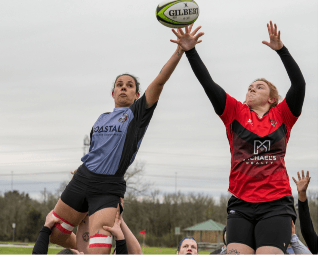 Two female rugby players jump to catch a ball during a game outdoors, with other players in the background.