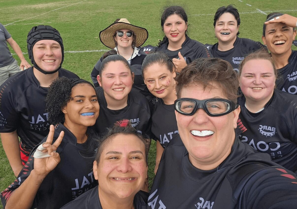 Group of smiling women and girls in sports uniforms outdoors, some making peace signs, on a grassy field.