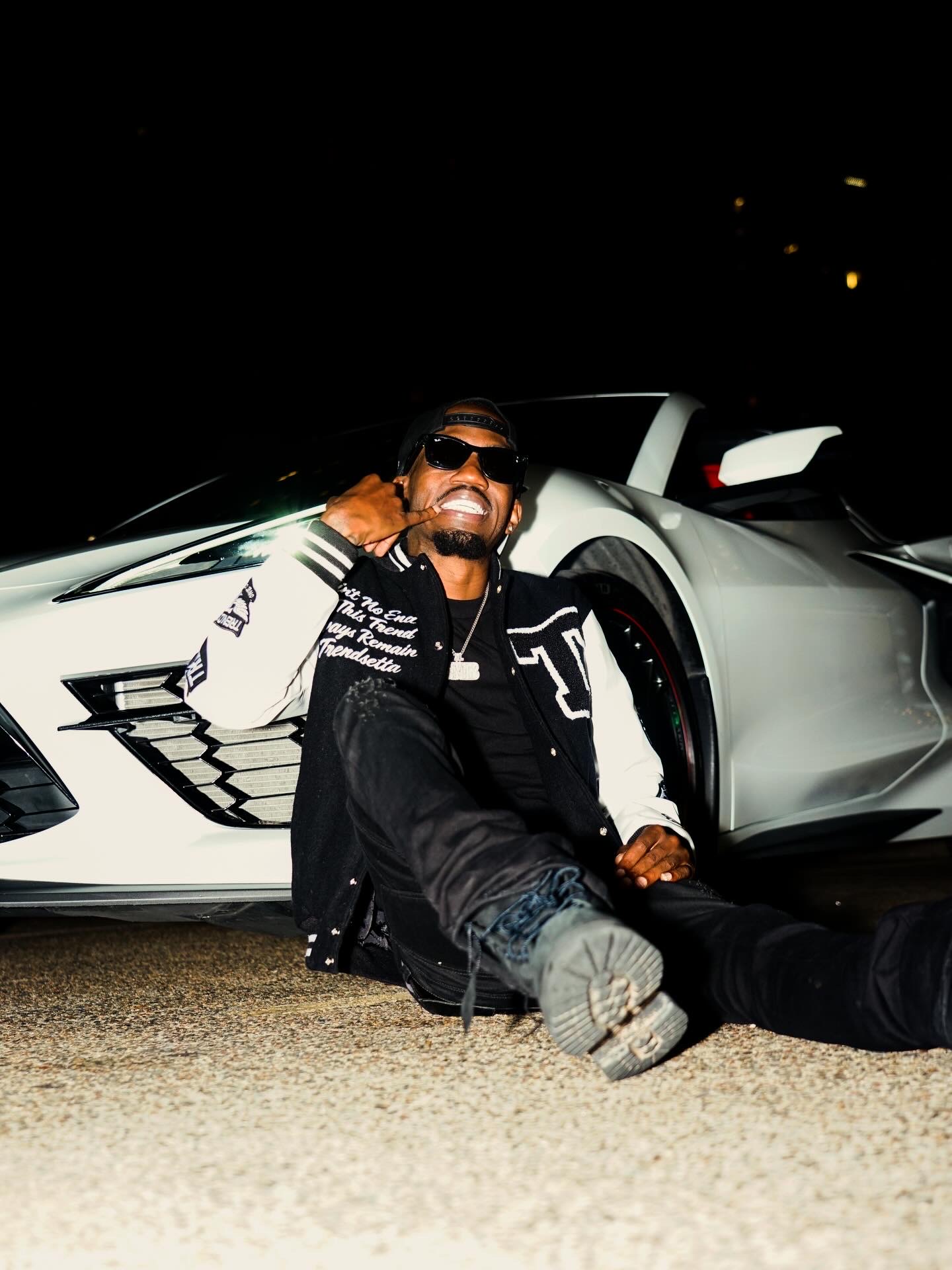 Person sitting on ground in front of a sports car at night, smiling and wearing sunglasses.