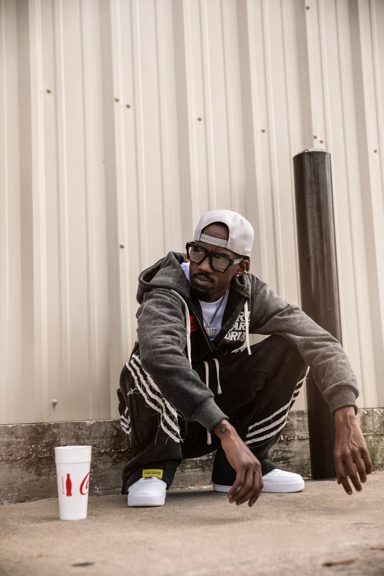 Person crouching in front of a metal wall, wearing a gray hoodie, white cap, glasses, and sneakers, with a drink cup nearby.