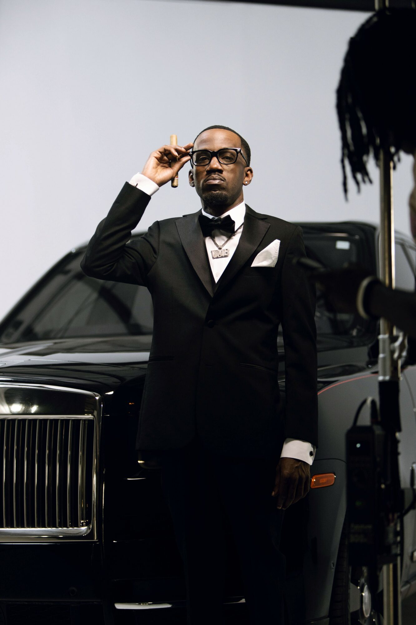 Man in tuxedo adjusting glasses, standing in front of a black luxury car, with a person partially visible on the right.