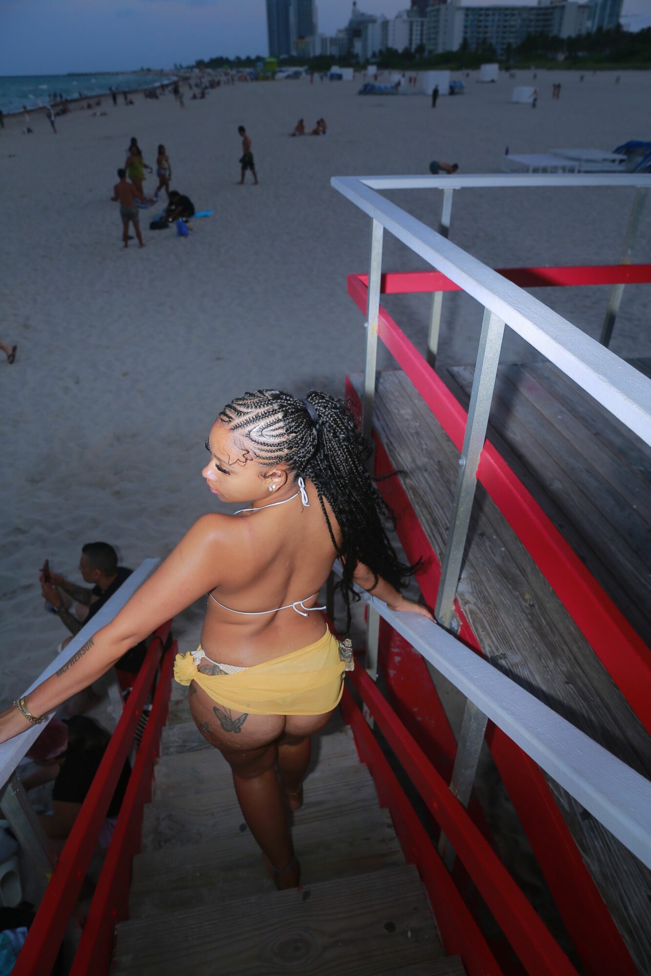 Woman in yellow shorts and sunglasses leaning on a red railing near a beach, with people in the background.