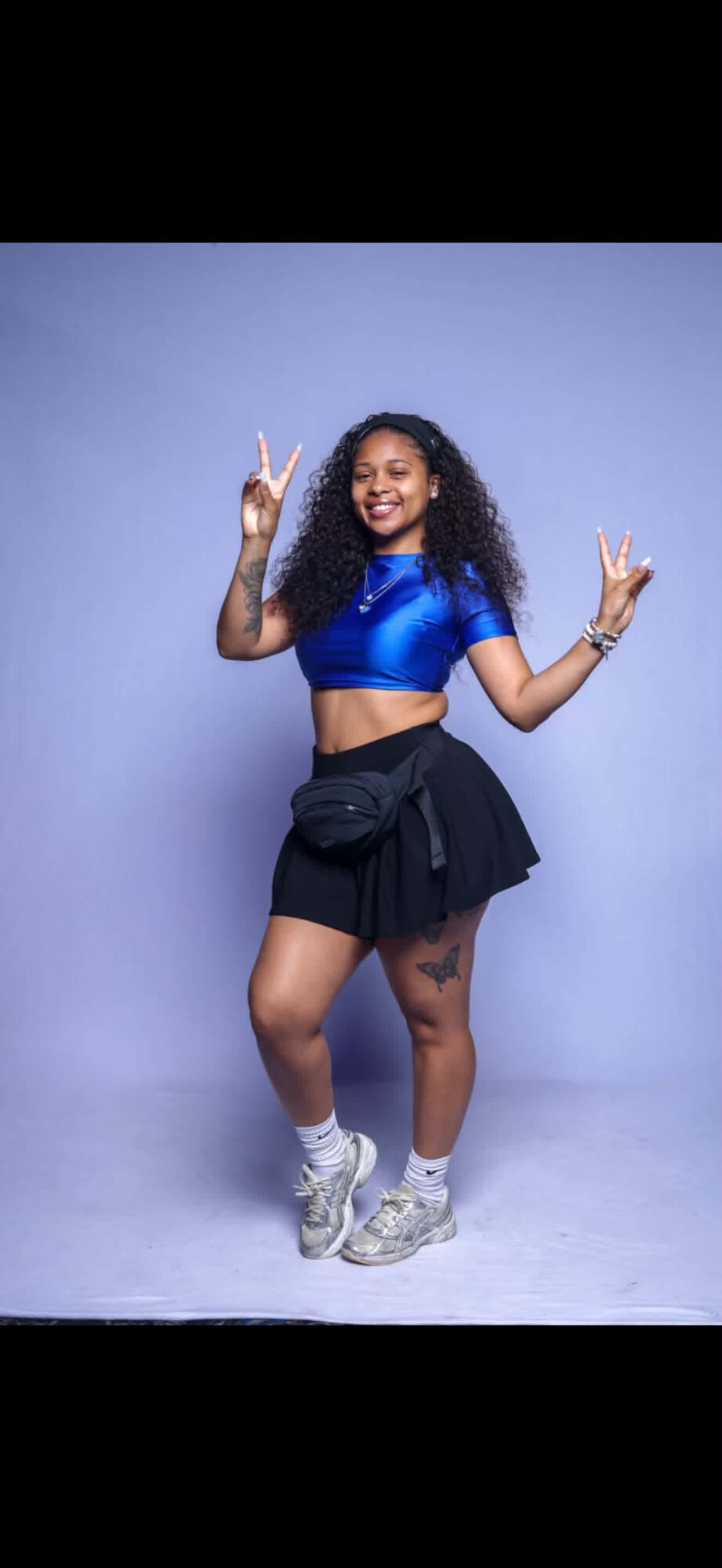 Young woman with curly hair in a blue crop top and black skirt, smiling and making peace signs, standing against a light purple background.