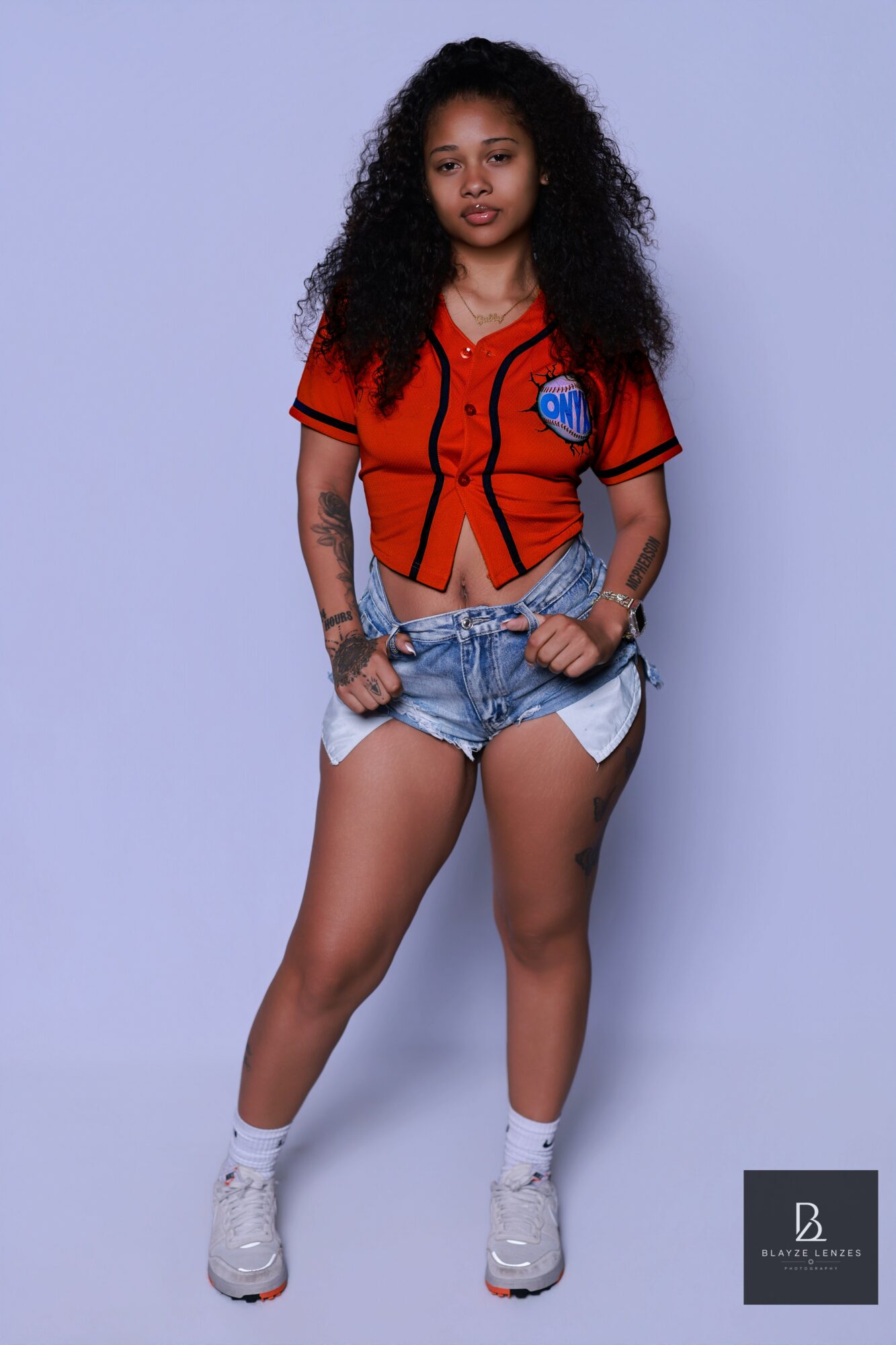 Young woman with curly hair wearing a red and black sports jersey, denim shorts, and white sneakers, standing against a light background.