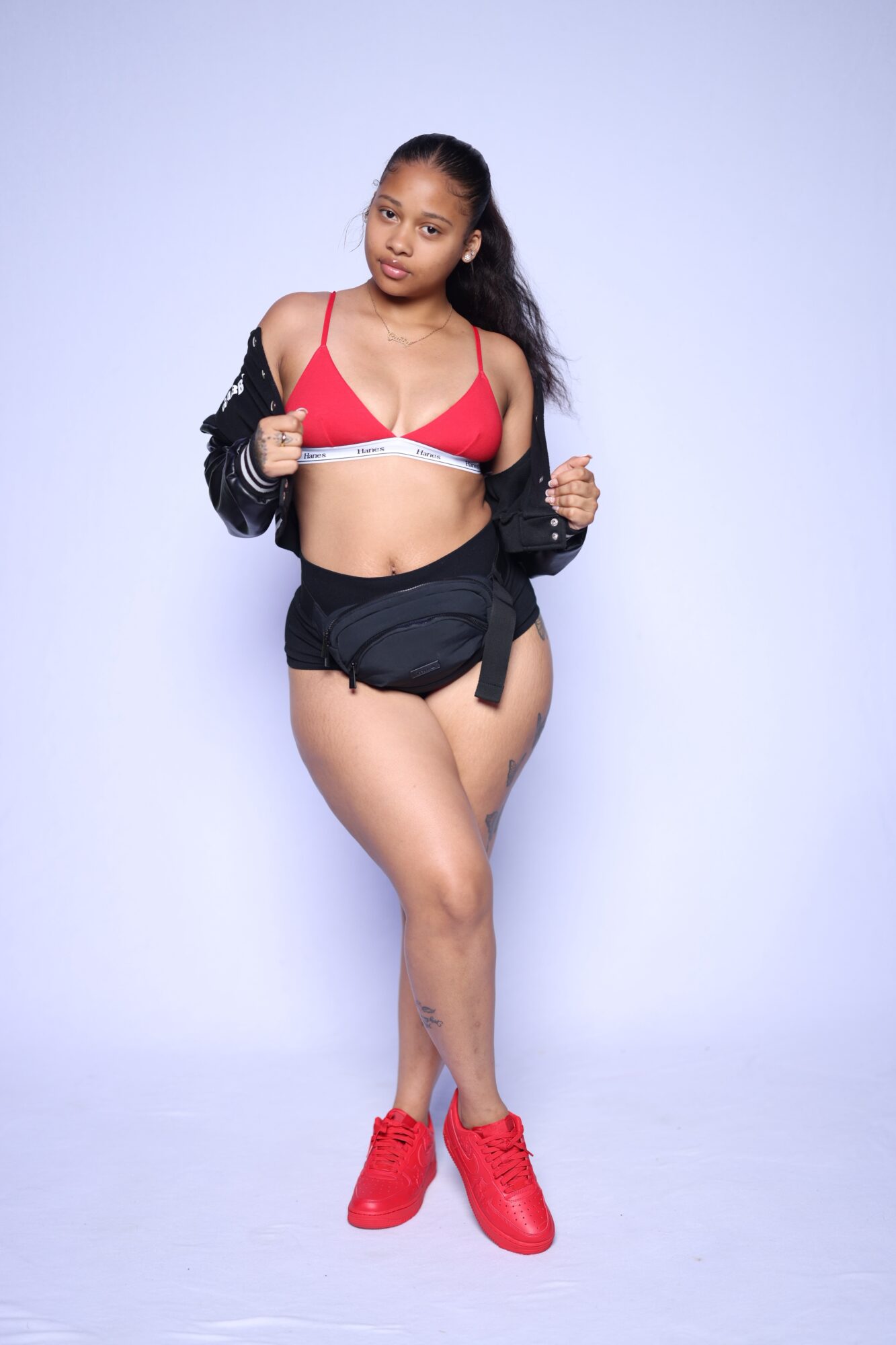 Young woman in red and white sports bra, black jacket, black shorts, and red sneakers standing against a light background.