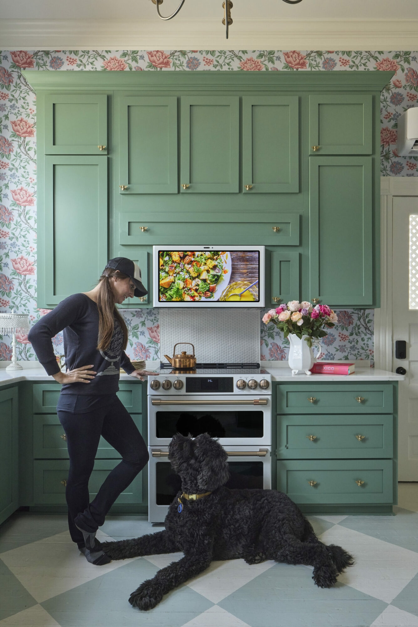 Woman standing in a kitchen with green cabinets, floral wallpaper, and a large black dog lying on the floor, flowers on the counter.