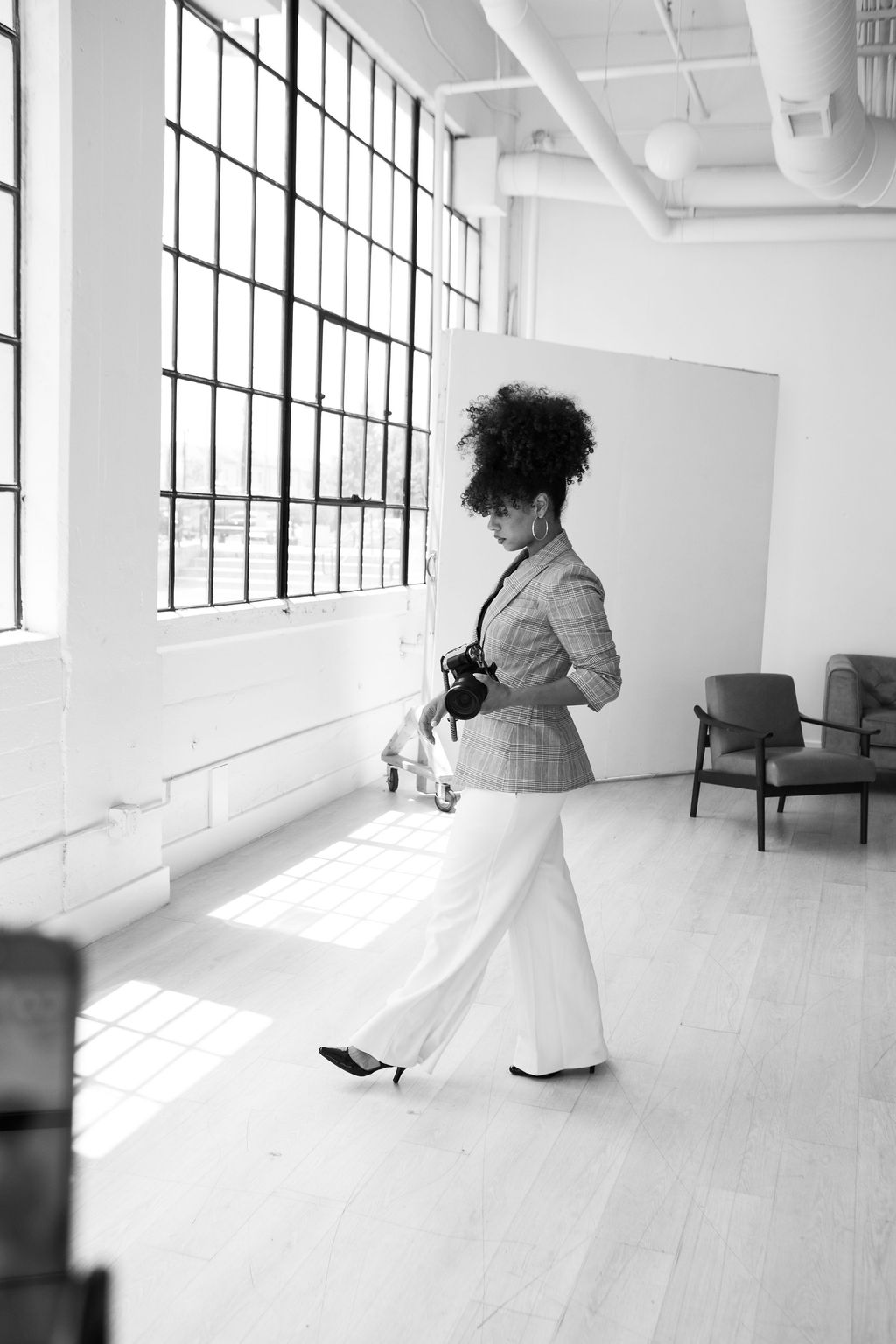 Woman with curly hair, wearing a blazer and wide-leg pants, walking in a bright, spacious room with large windows.