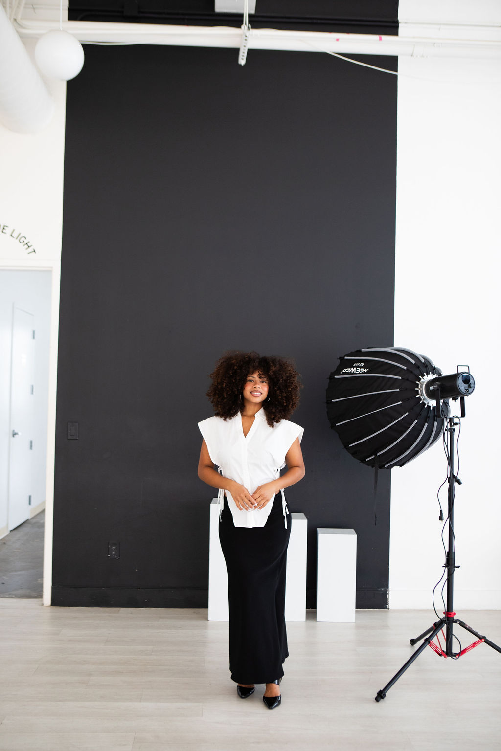 Woman with curly hair in white top and black pants stands in a photography studio with a large black backdrop and professional lighting equipment.
