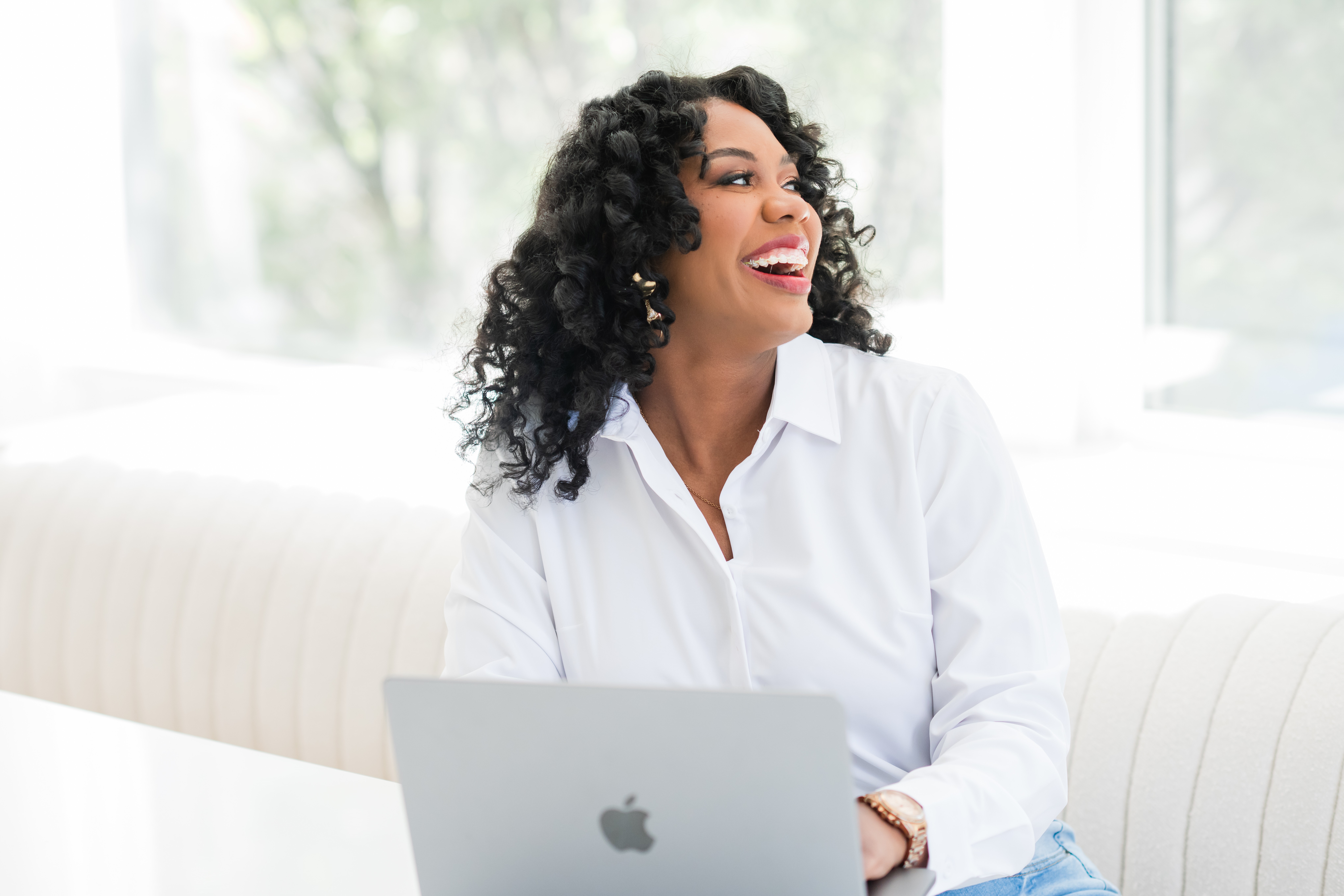 Woman with curly hair smiling, sitting at a table with a laptop, in a bright room with large windows.