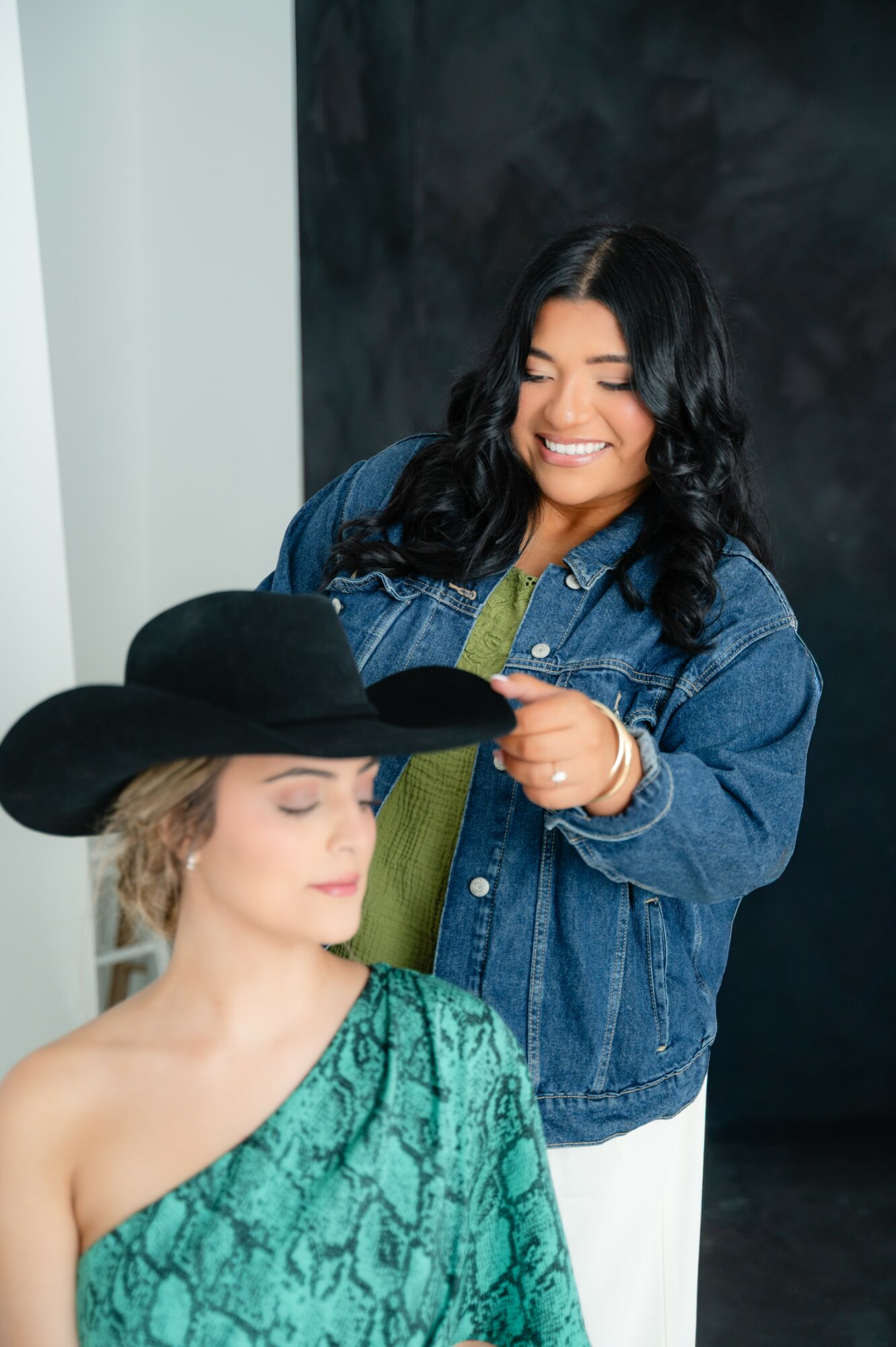 Two women, one smiling and holding a hat, the other wearing a hat and looking down, in a studio setting.
