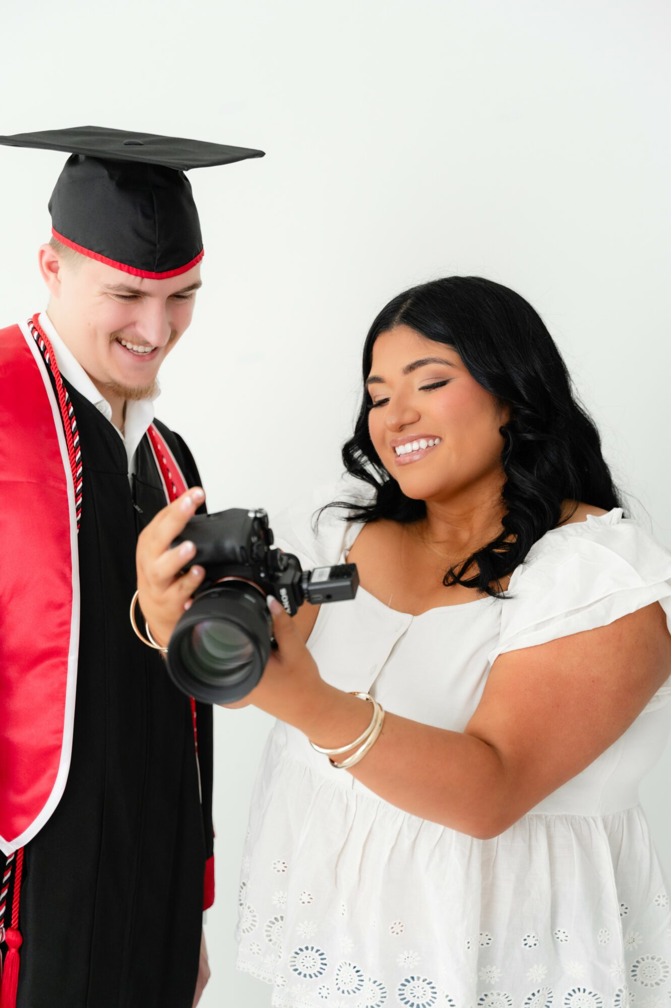 Two people, one in graduation attire and the other in a white dress, look at a camera together, smiling.