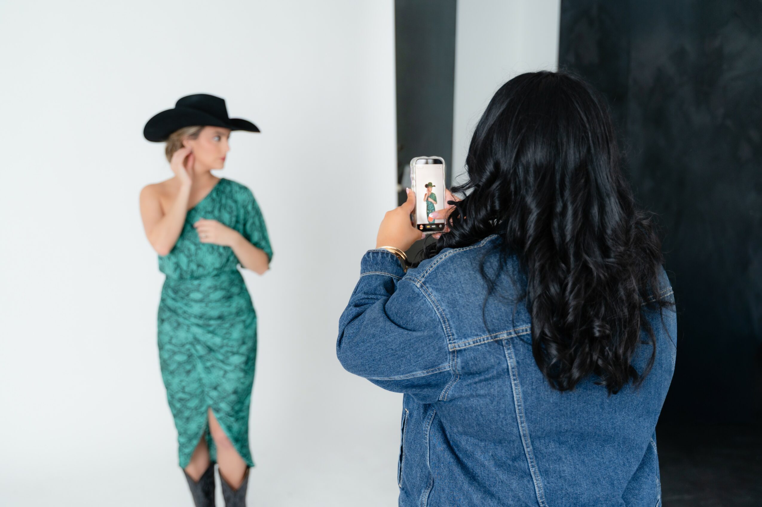 Person with long dark hair taking a photo of a woman in a green dress and black hat, standing against a white wall.