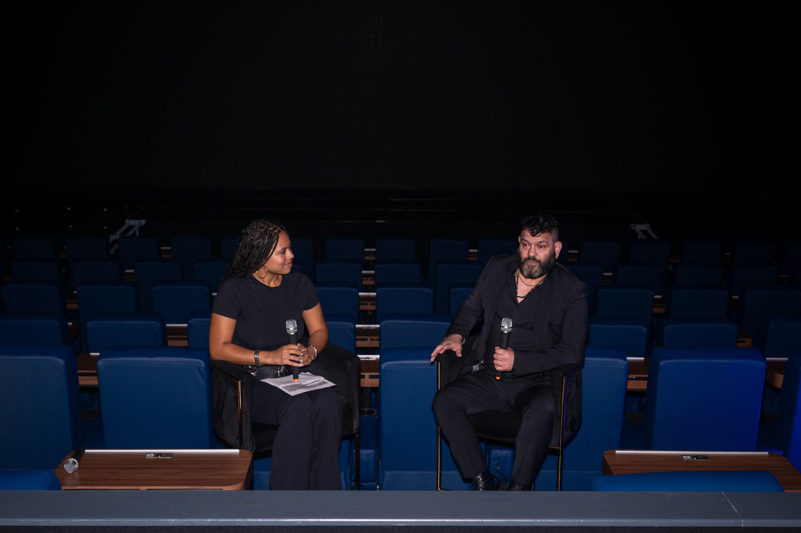 Two people sit on stage with microphones, engaged in conversation, in an empty auditorium with blue seats behind them.