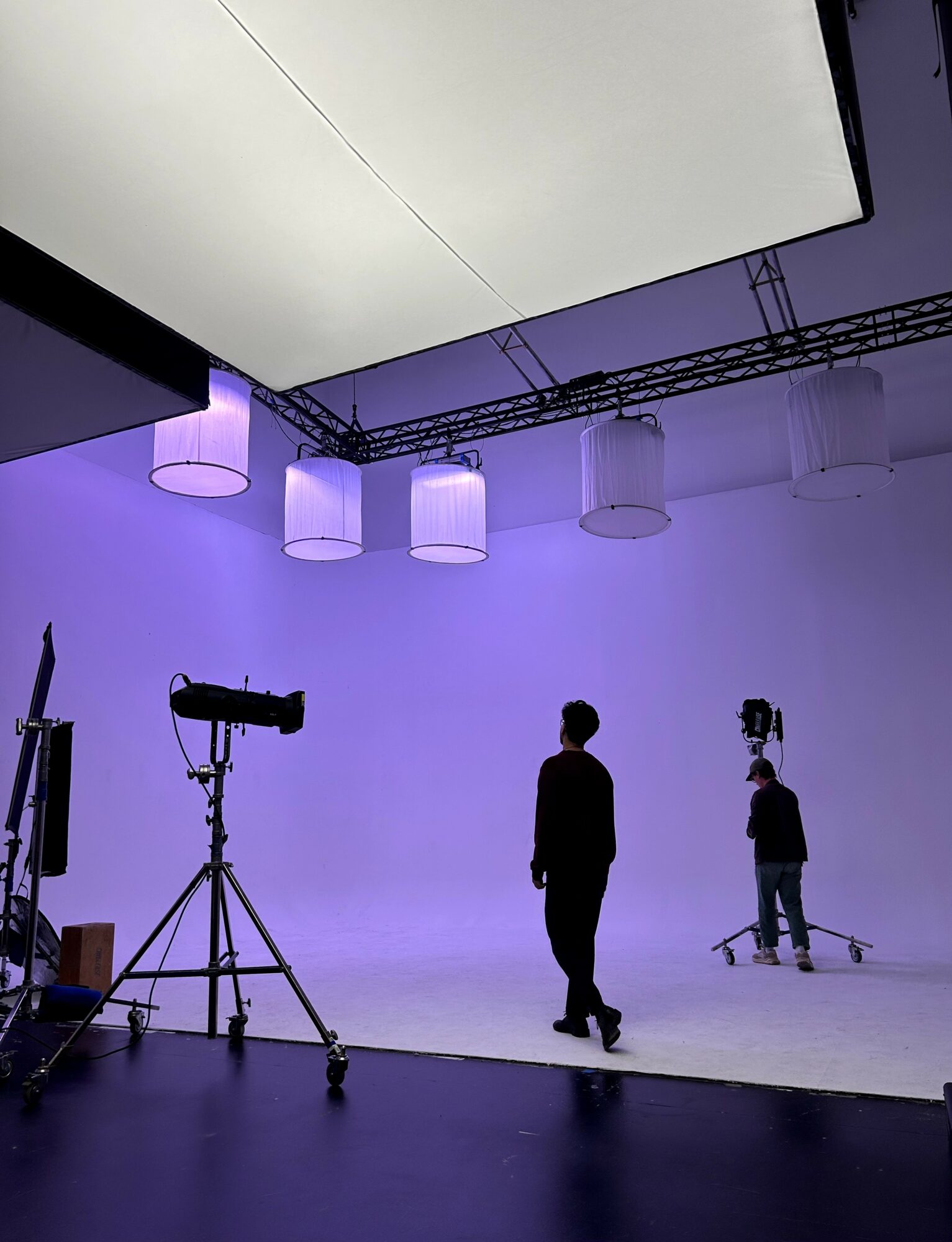 Person standing in a studio with purple lighting, camera equipment, and large hanging lights on ceiling.