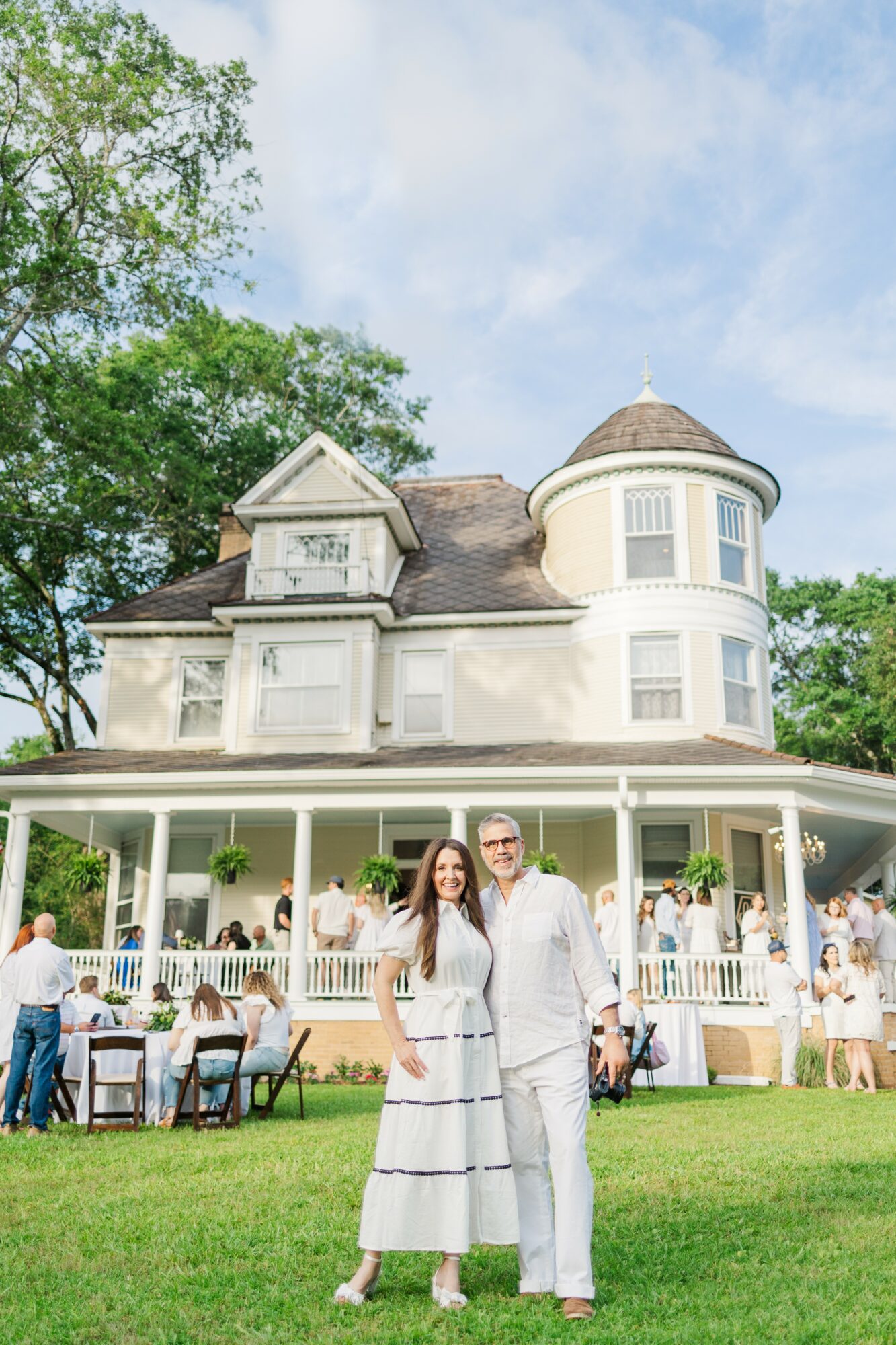 A couple stands on a lawn in front of a large white house with a porch, with people dining outside. Bright sky.