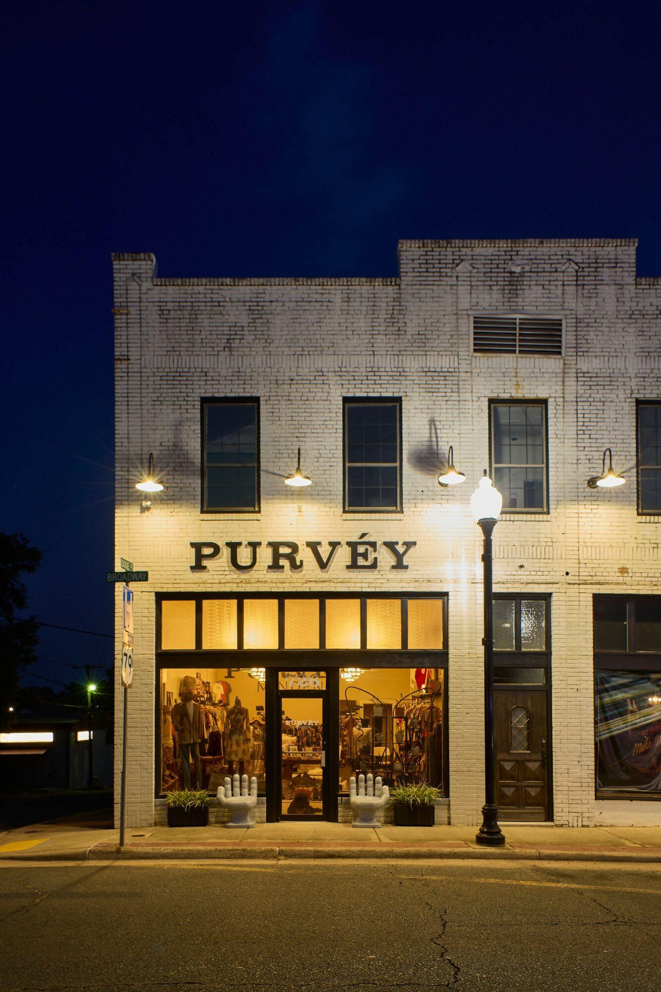 Storefront with illuminated sign reading 'PURVÉY' and large glass windows showing interior displays, on a city street at night.