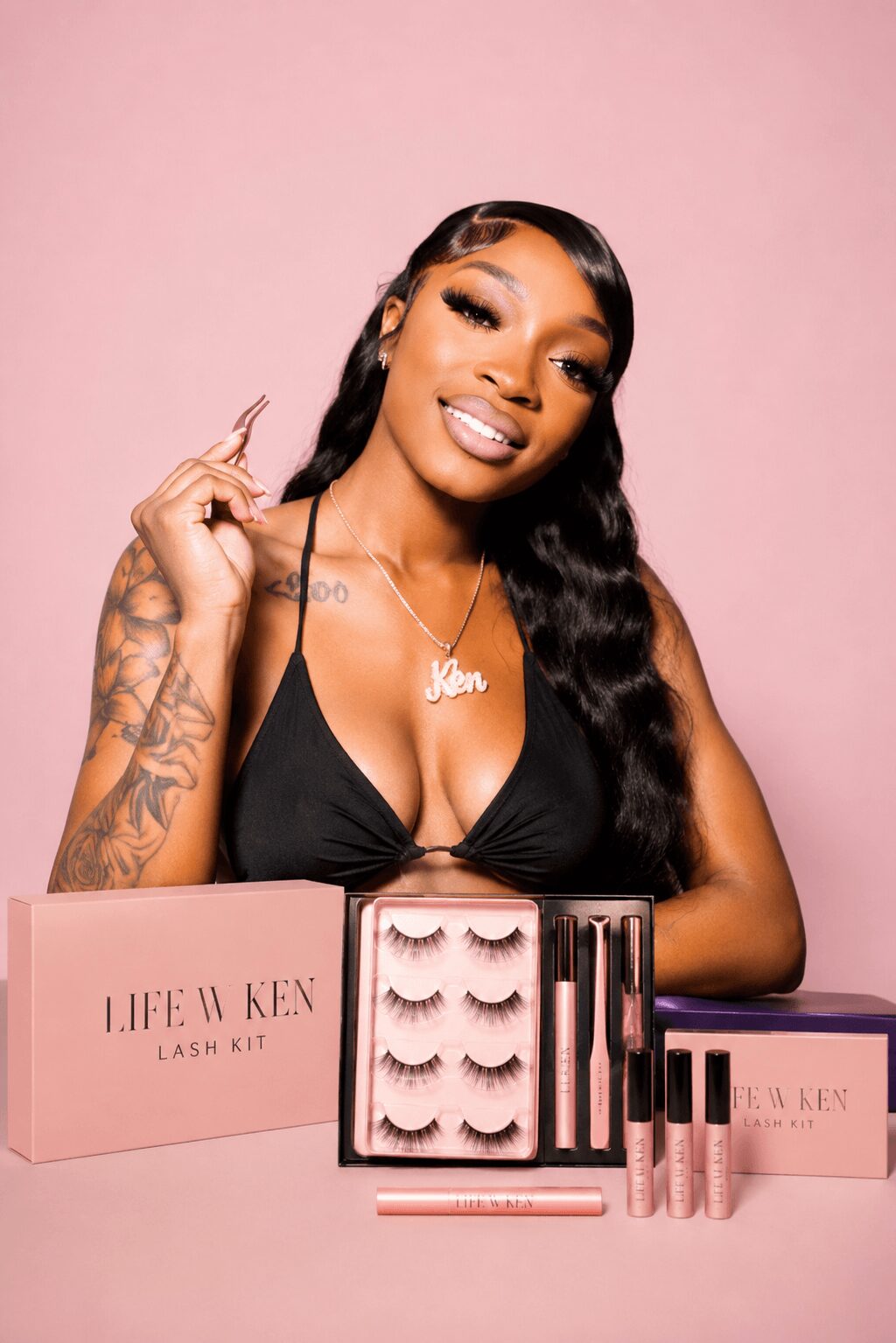 Smiling woman with long wavy hair and tattoos displays eyelash products against pink background.