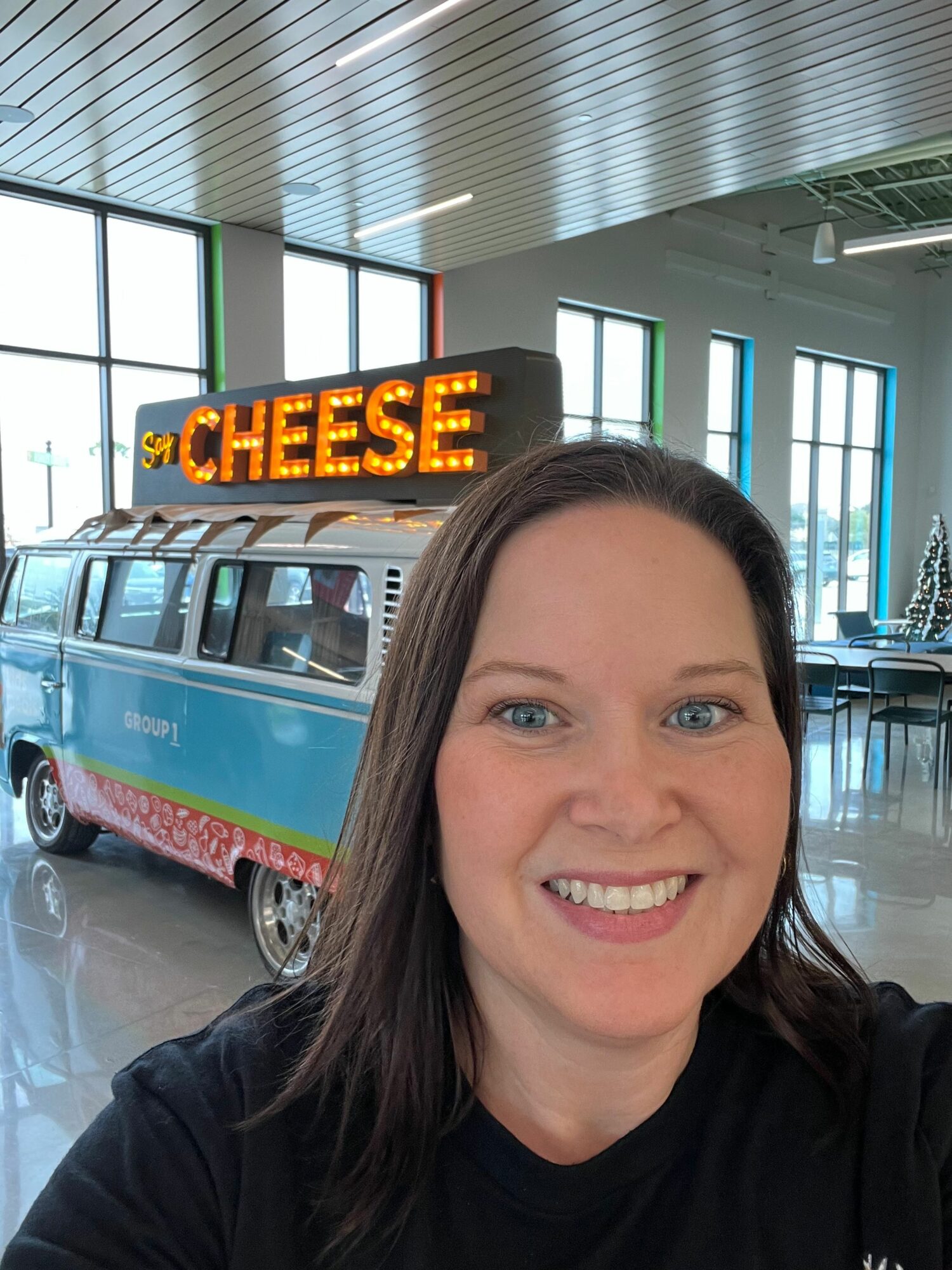 Smiling woman with dark hair in a black shirt takes a selfie inside a building with large windows and a sign that says 'CHEESE'.