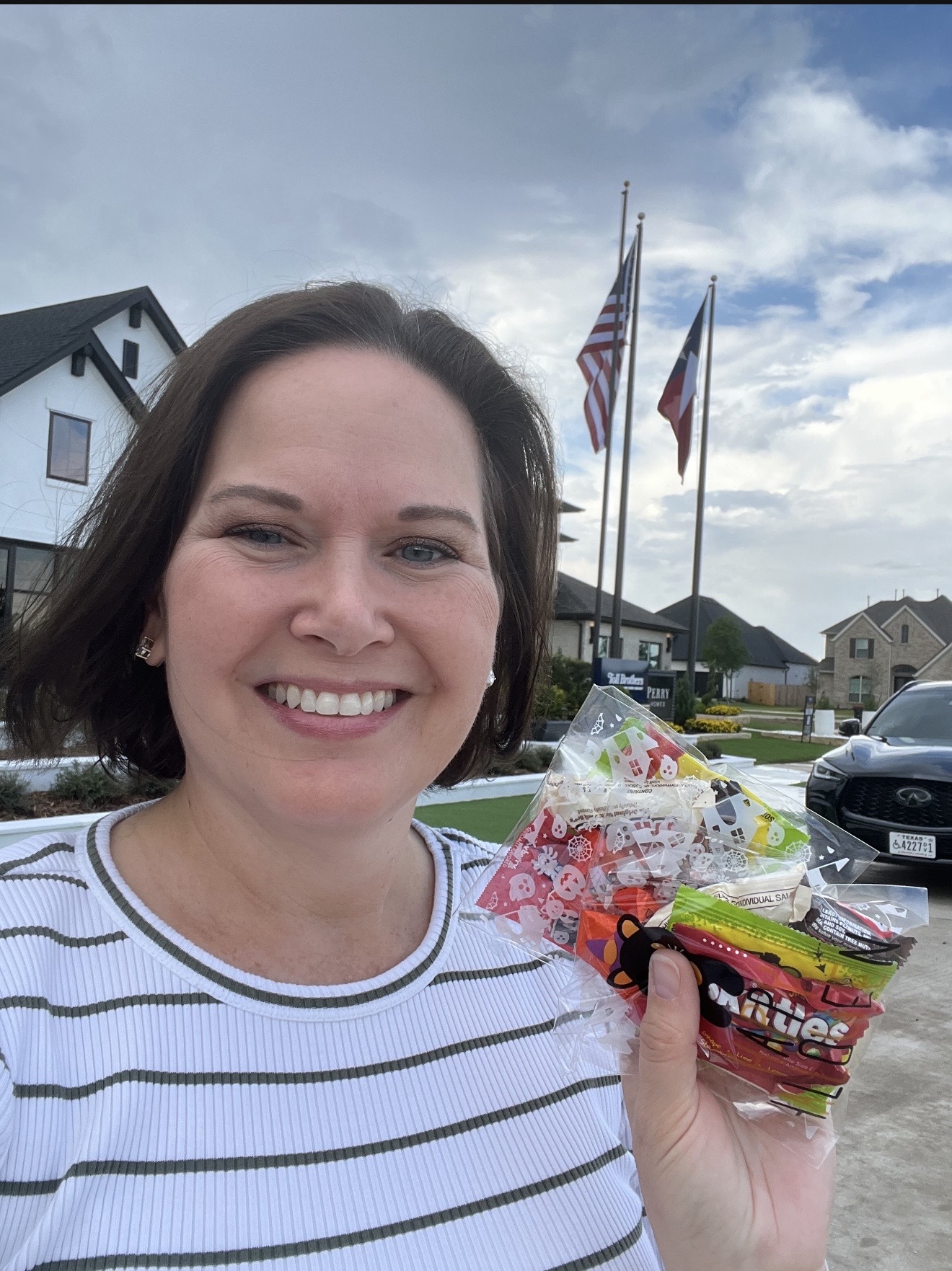 Woman smiling outdoors holding a bag of candy, with houses, flags, and cars in the background.