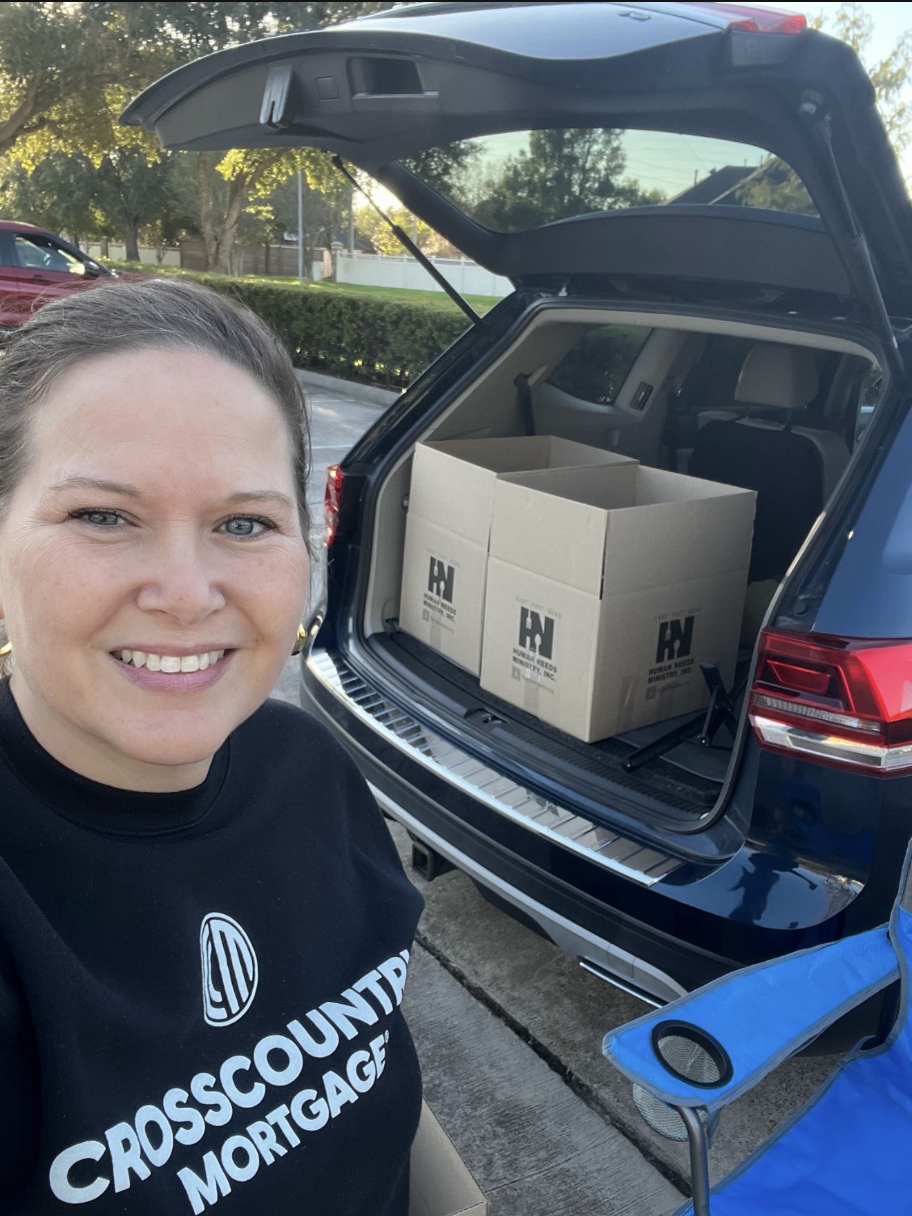 Woman smiling in front of open car trunk with boxes inside, parked outdoors with trees and sunlight.