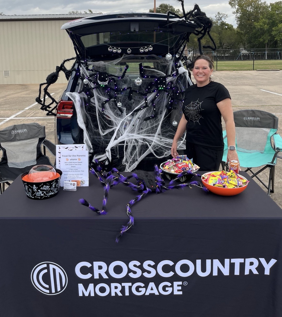 Person standing next to a decorated vehicle with spider web decorations, at a table with candy bowls, outdoors.