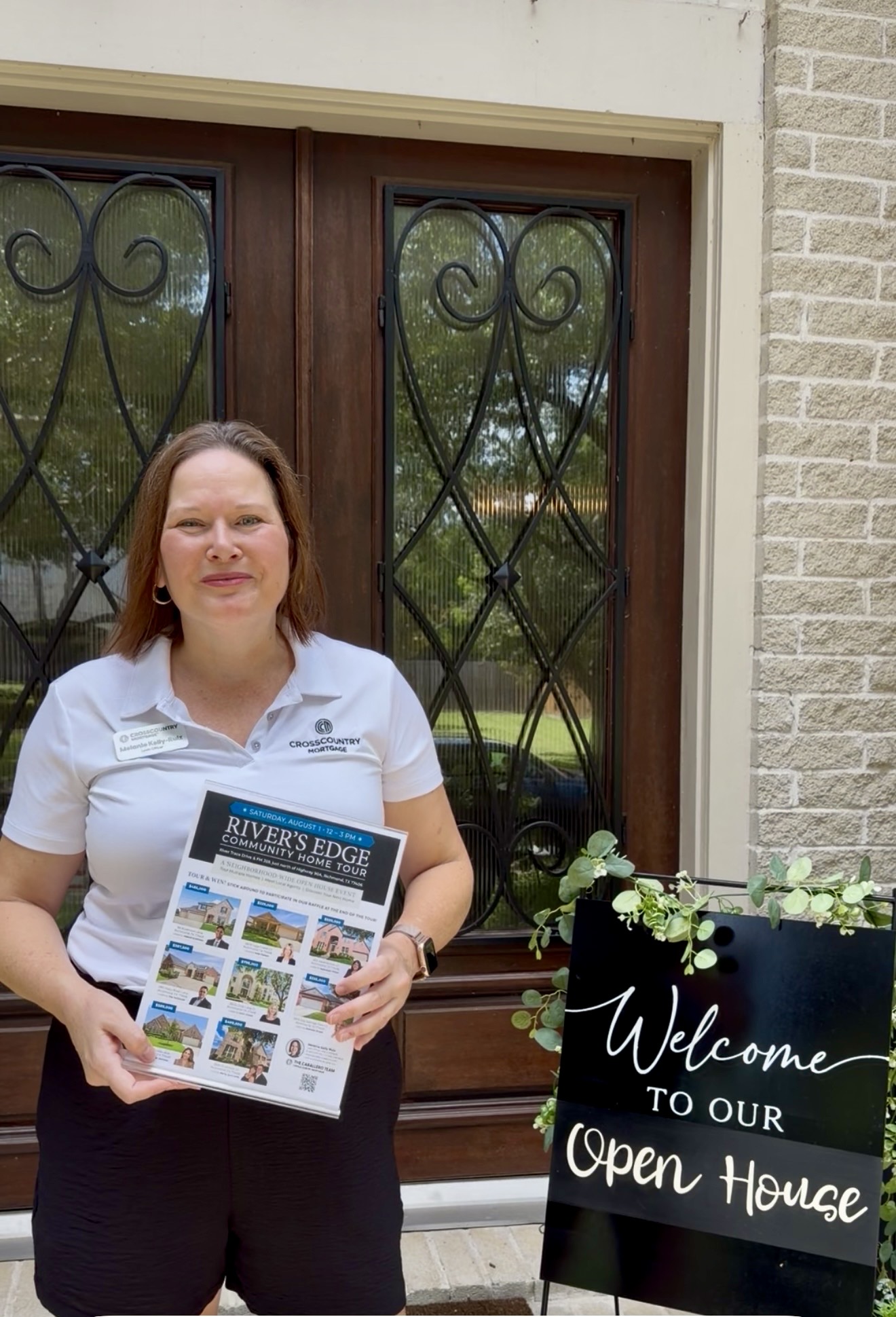 Woman holding a flyer standing outside a building with a welcome sign, brick wall, and decorative window. Worded sign reads 'Welcome to our Open House'.