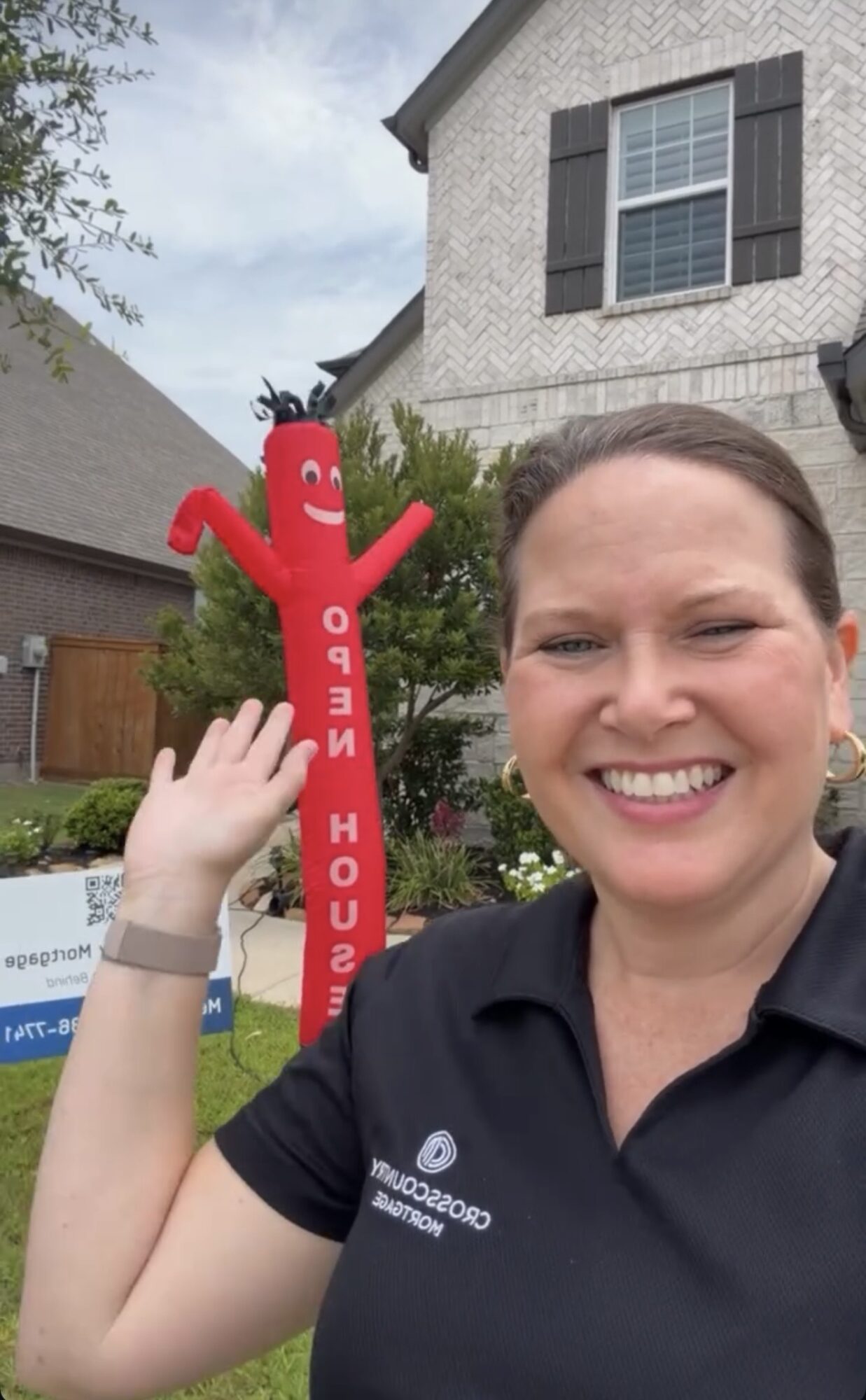 Woman smiling outdoors with a red inflatable character in the background, house and trees visible.