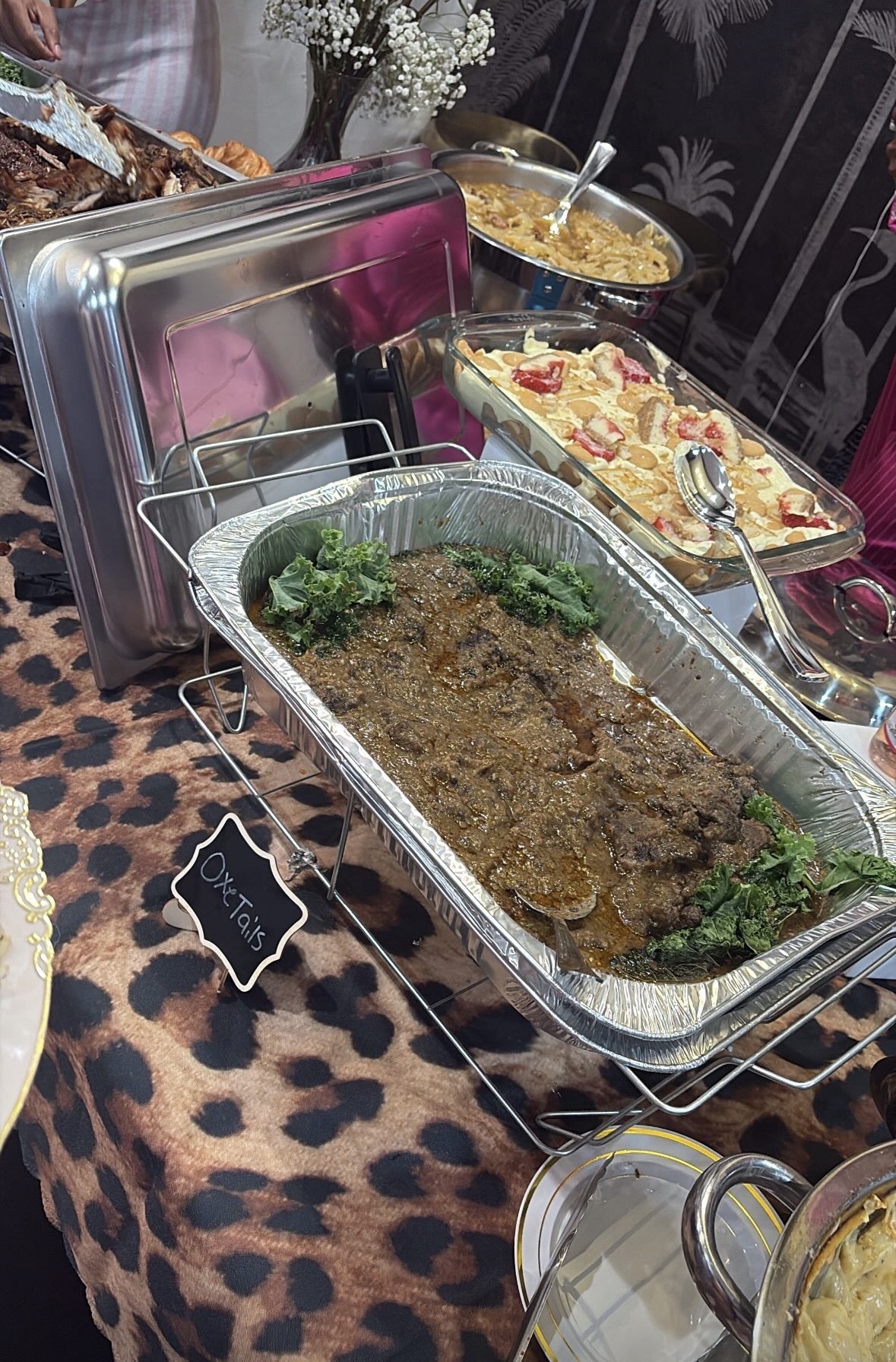 Assorted dishes on a table with a leopard print tablecloth, including a large tray of cooked meat garnished with parsley, a casserole, and a bowl of soup.