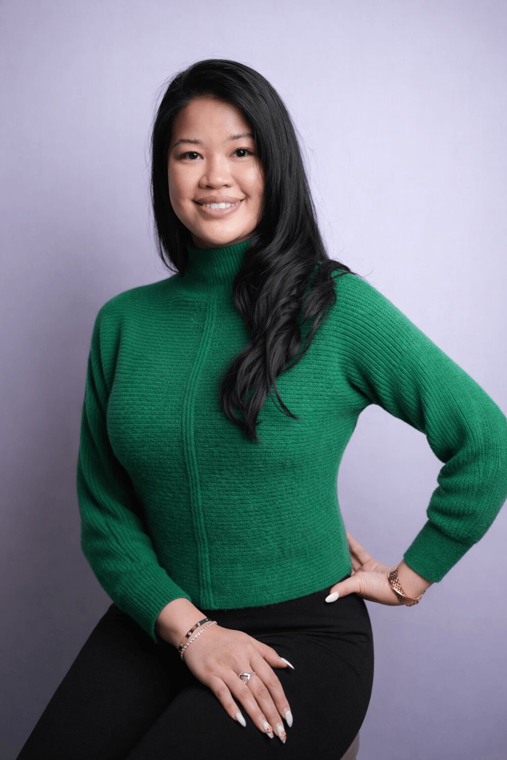 Woman with long black hair wearing a green sweater and black pants, smiling, standing against a light background.