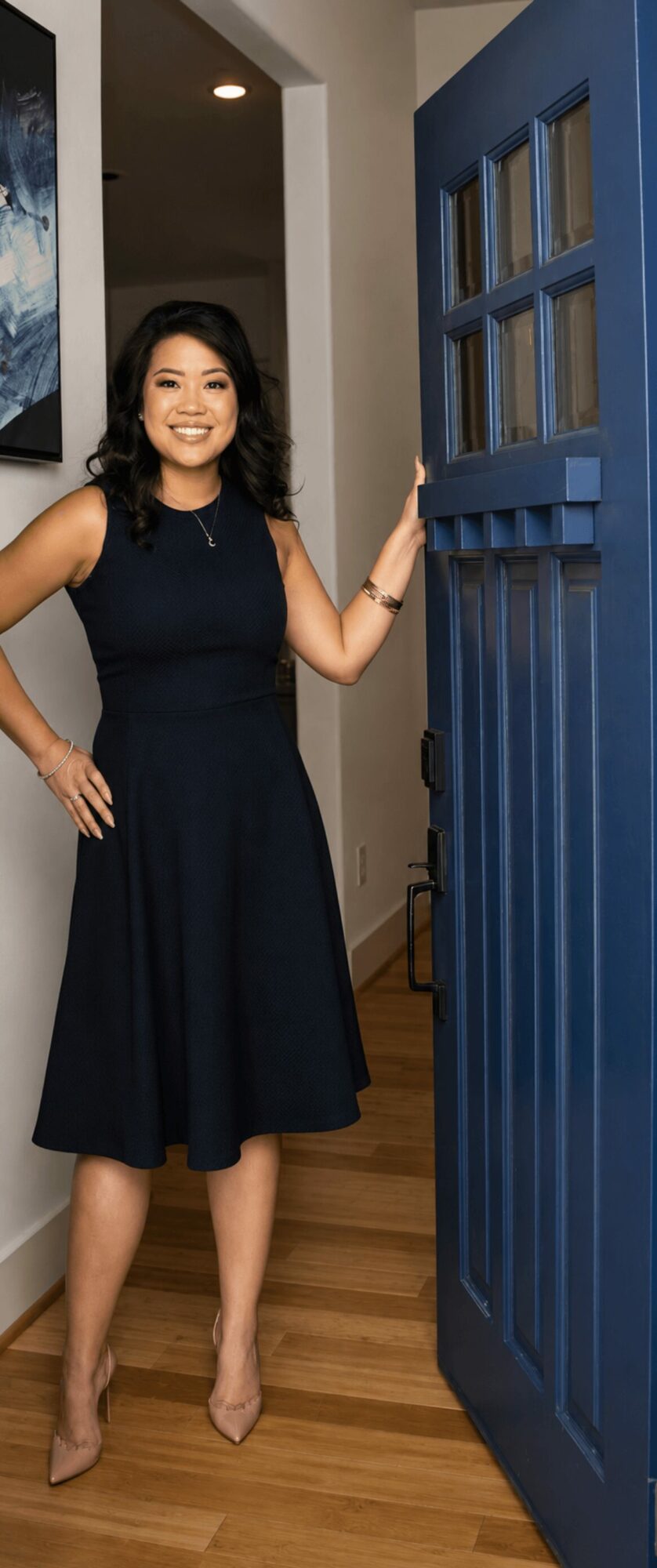 Woman standing in doorway, smiling, wearing a dark dress, with a blue door and wooden floor visible.