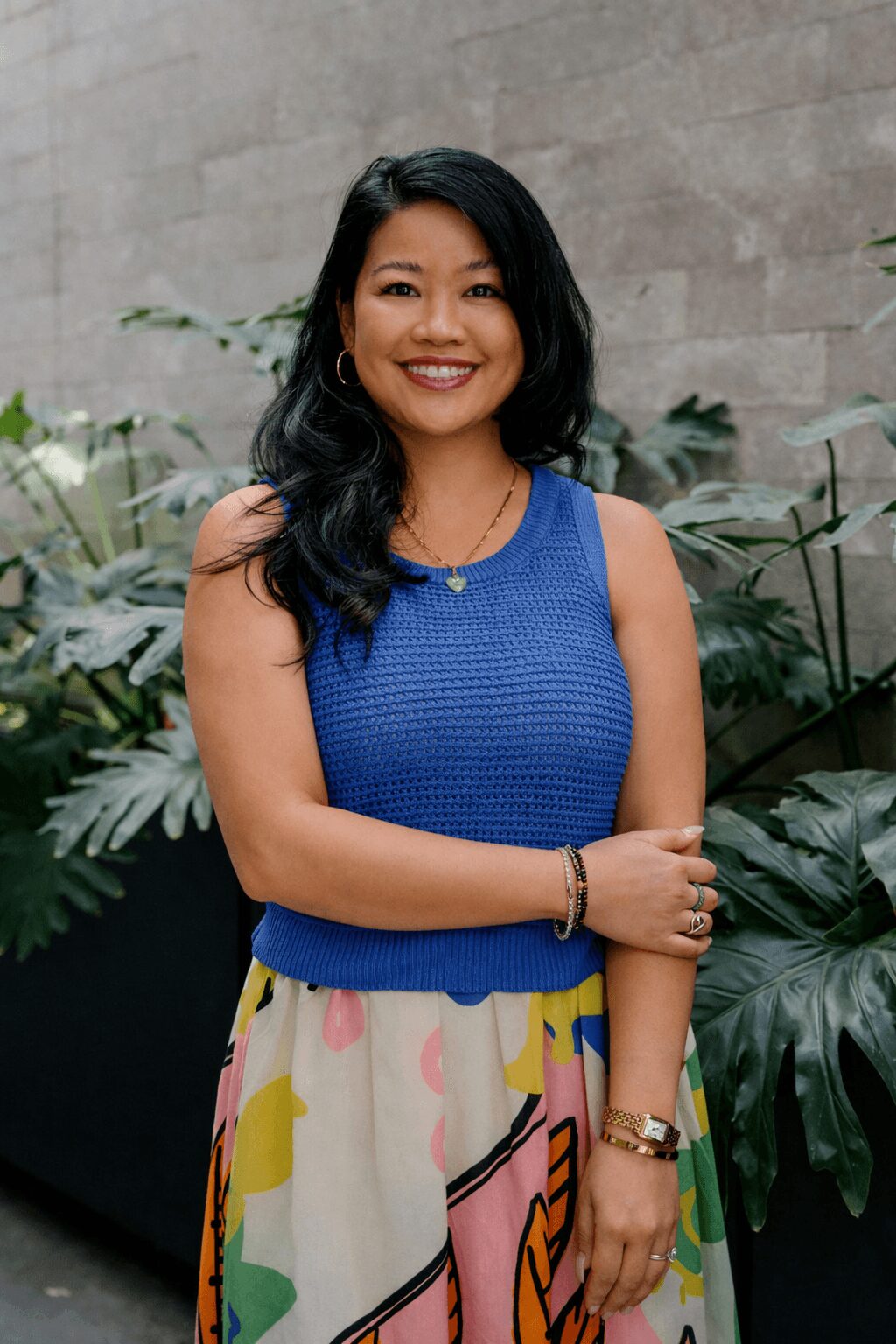 Woman with long black hair smiling, wearing a blue sleeveless top and a colorful skirt, standing outdoors with plants.